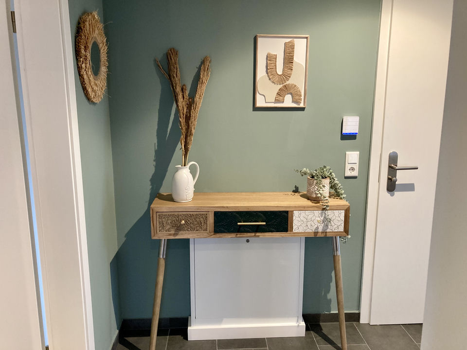 Angolo d’ingresso con un elegante tavolo consolle in legno con tre cassetti decorati, una brocca bianca con fiori secchi, un’opera in rafia incorniciata e uno specchio rotondo in paglia su una parete verde salvia.