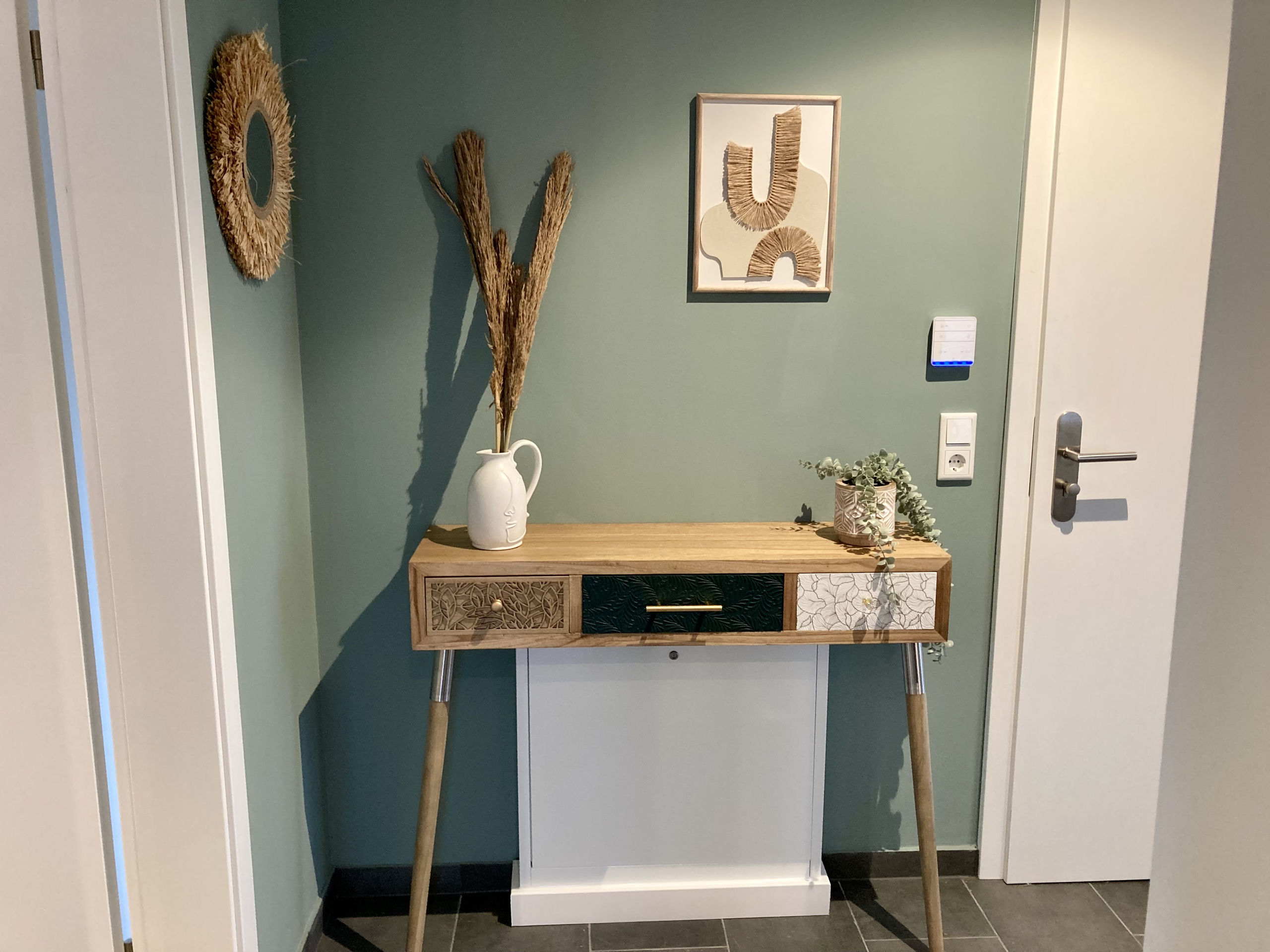 Eingangsecke mit schlankem Holz-Konsolentisch und drei gemusterten Schubladen, weißer Kanne mit Trockengräsern, gerahmtem Raffia-Bild und rundem Strohspiegel an salbeigrüner Wand.