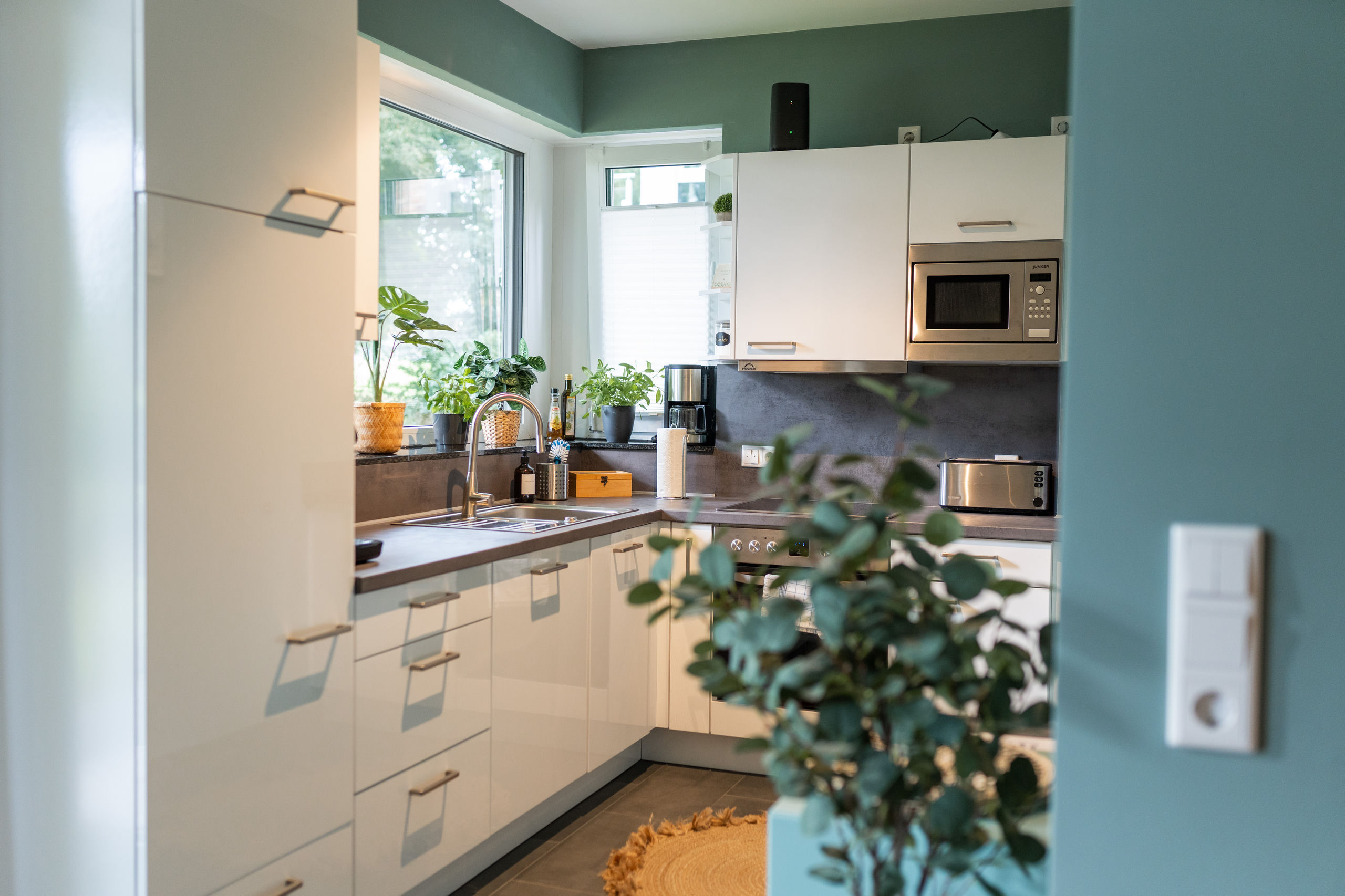 Kompakte Küche mit hochglänzenden weißen Schränken, dunklen Arbeitsplatten, einer Fensterbank voller Topfpflanzen, Spüle mit hoher Armatur, Einbaumikrowelle, Kaffeemaschine und Toaster sowie Induktionskochfeld mit Backofen.