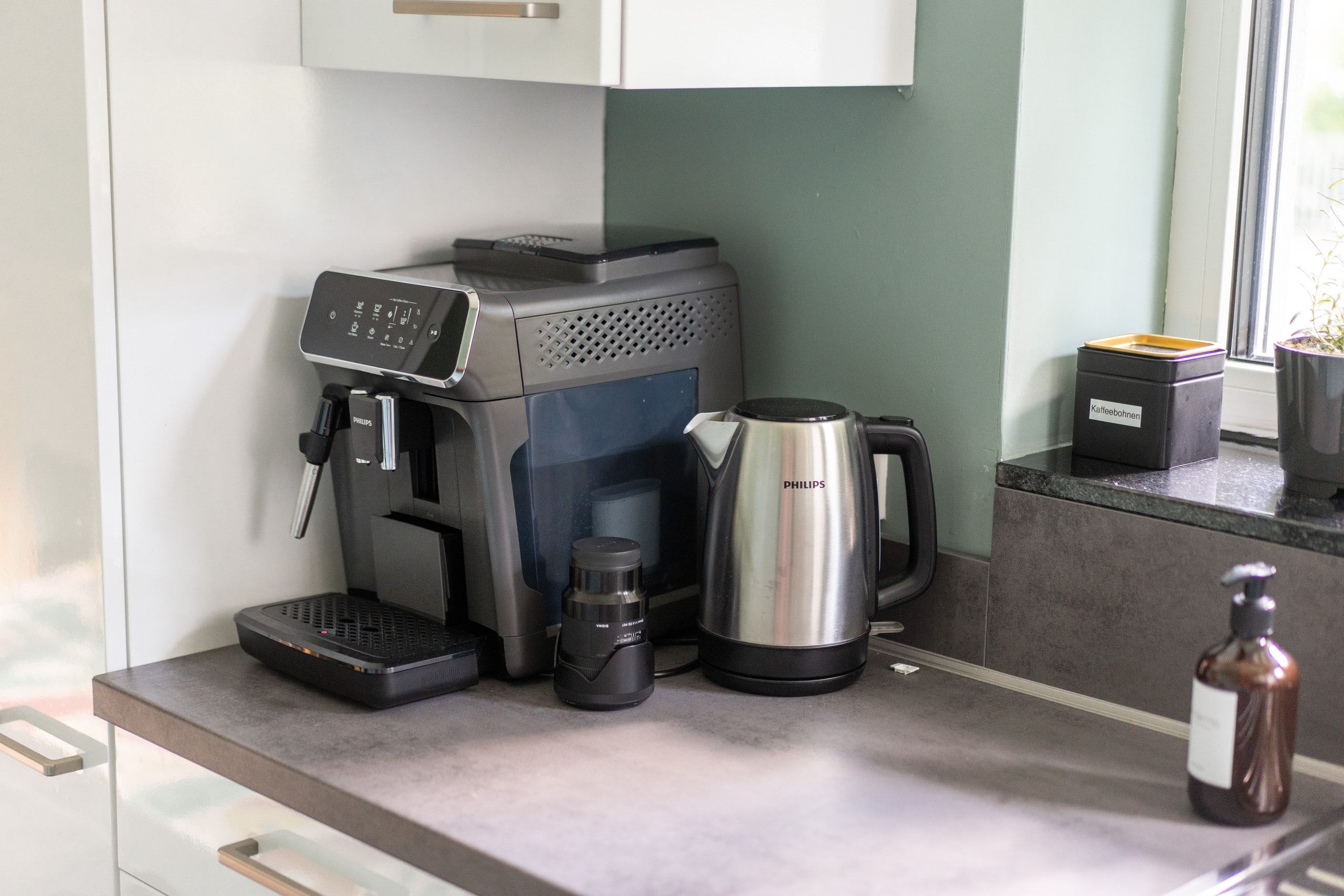 Kaffeeplatz auf der Arbeitsplatte mit vollautomatischer Espressomaschine mit Touchdisplay und Milchaufschäumer, einem Edelstahl-Wasserkocher und einer beschrifteten Dose mit Kaffeebohnen.