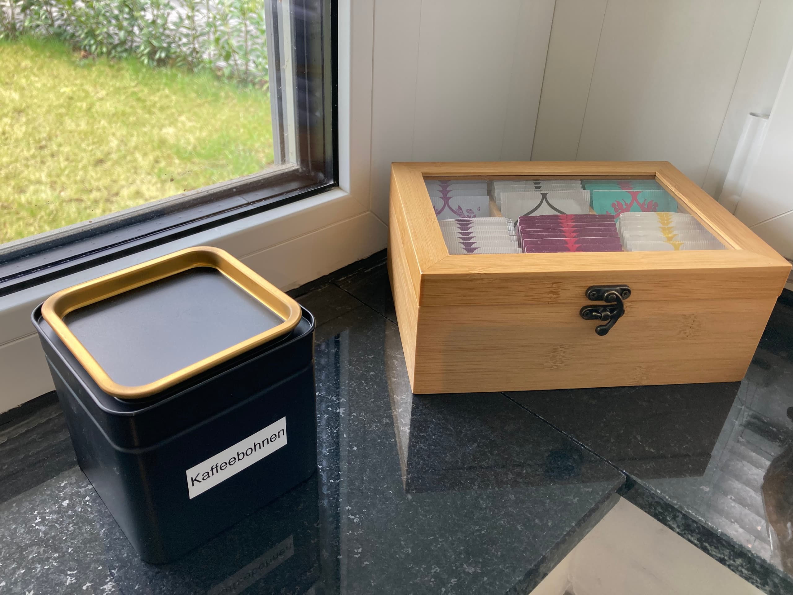 Auf dunkler Fensterbank eine schwarze Dose mit der Aufschrift „Kaffebohnen" neben einer Bambusbox mit Glasdeckel, gefüllt mit portionierten Kaffee- und Teebeuteln.