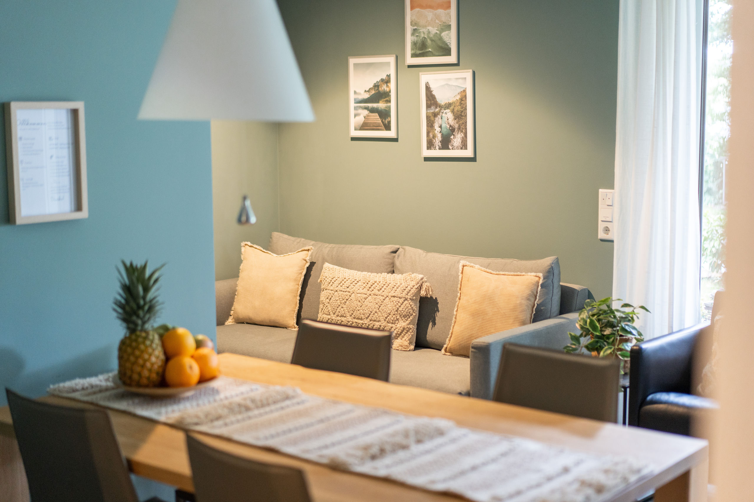Wohn- und Essbereich mit grauem Sofa in ockerfarbenen Kissen, drei gerahmten Naturmotiven an grüner Wand und einem Esstisch aus Eiche mit Obstschale im Vordergrund.