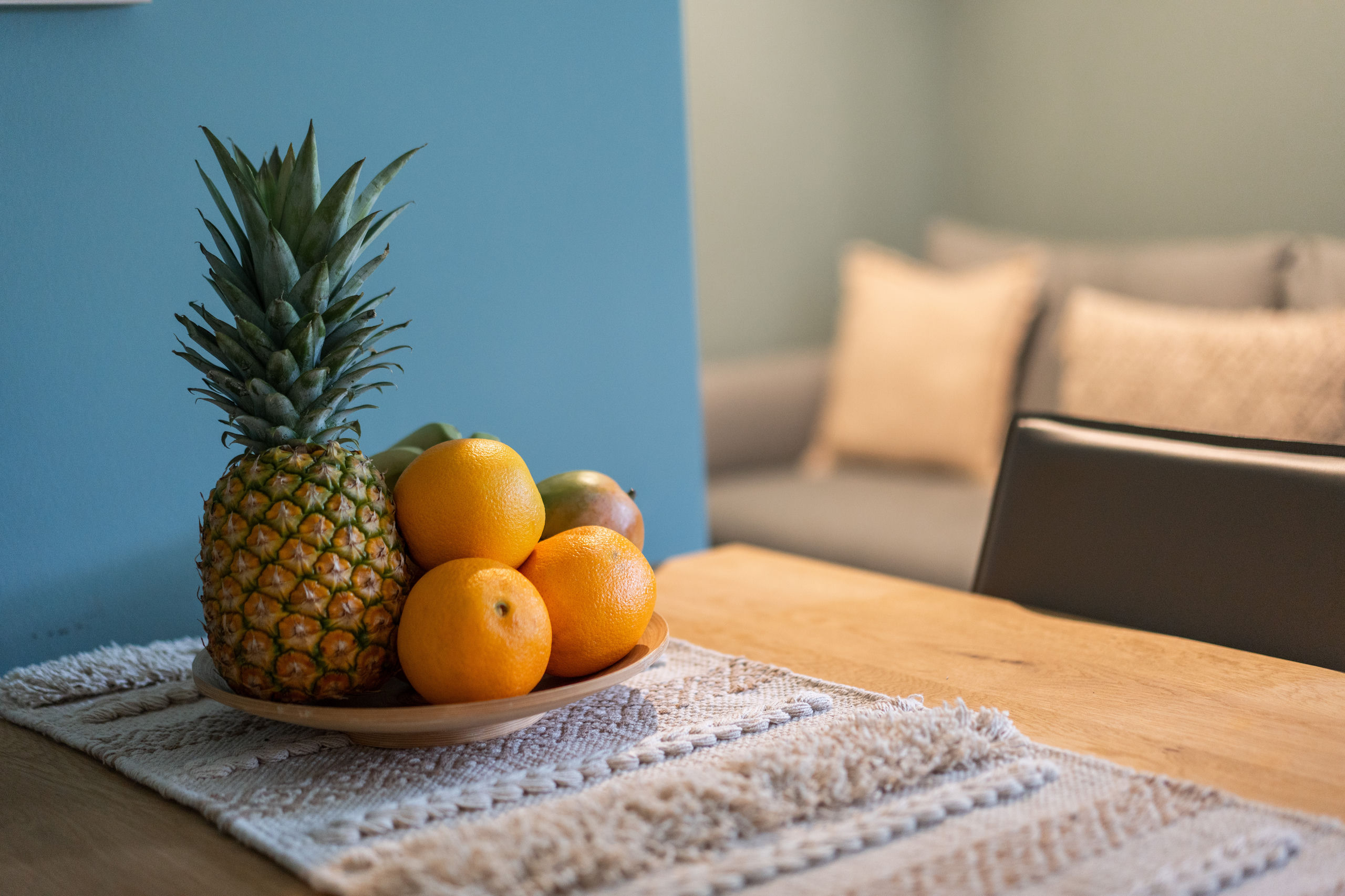 Nahaufnahme einer Holzschale mit Ananas, Orangen und Mango auf einem cremefarbenen, gewebten Tischläufer, dahinter unscharf das Sofa.