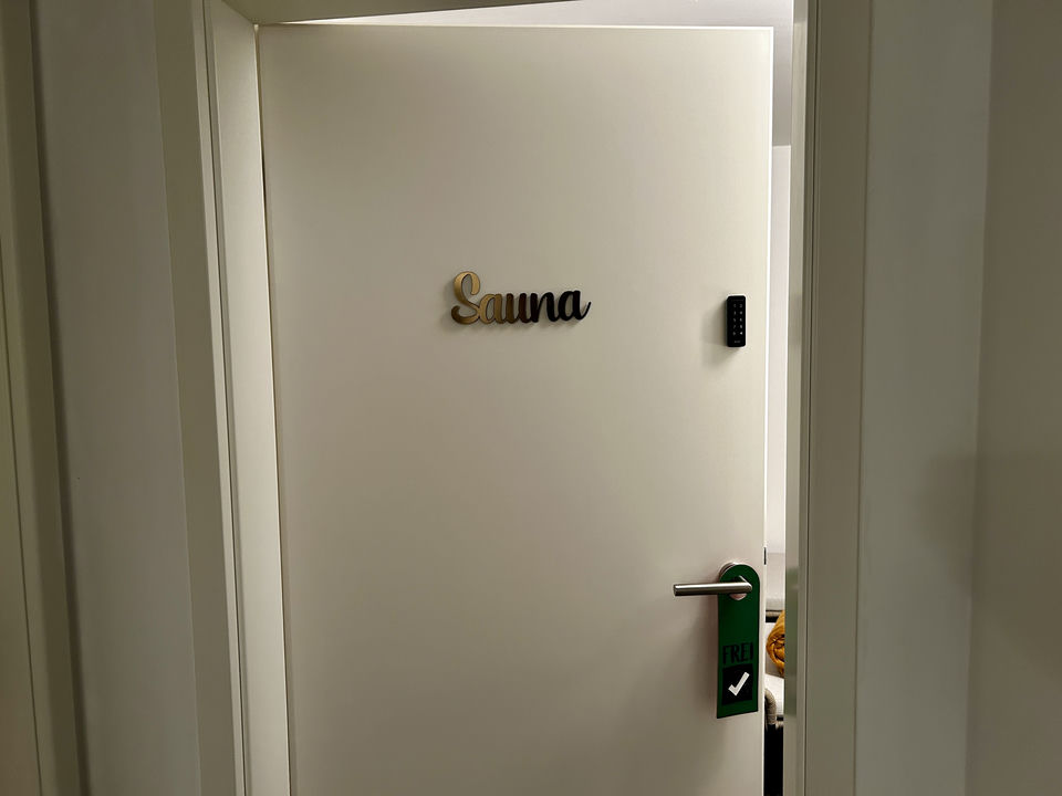 Una porta bianca leggermente socchiusa nell’ingresso reca un’insegna in legno con la scritta 'Sauna' e un’etichetta verde FREI appesa alla maniglia.