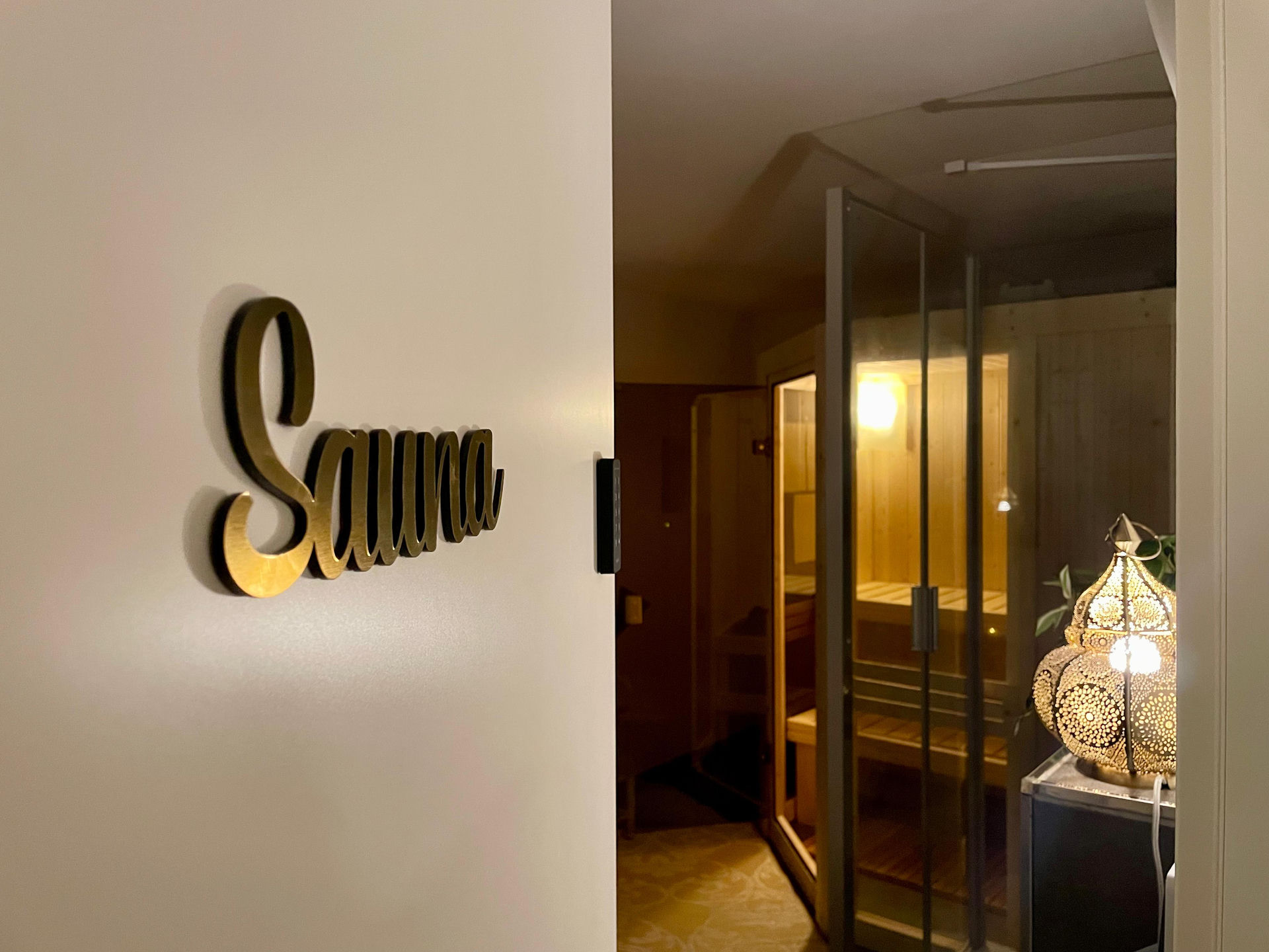 Gold Sauna lettering on a pale wall beside a doorway, with the glowing glass sauna cabin and a pierced brass lantern beyond.