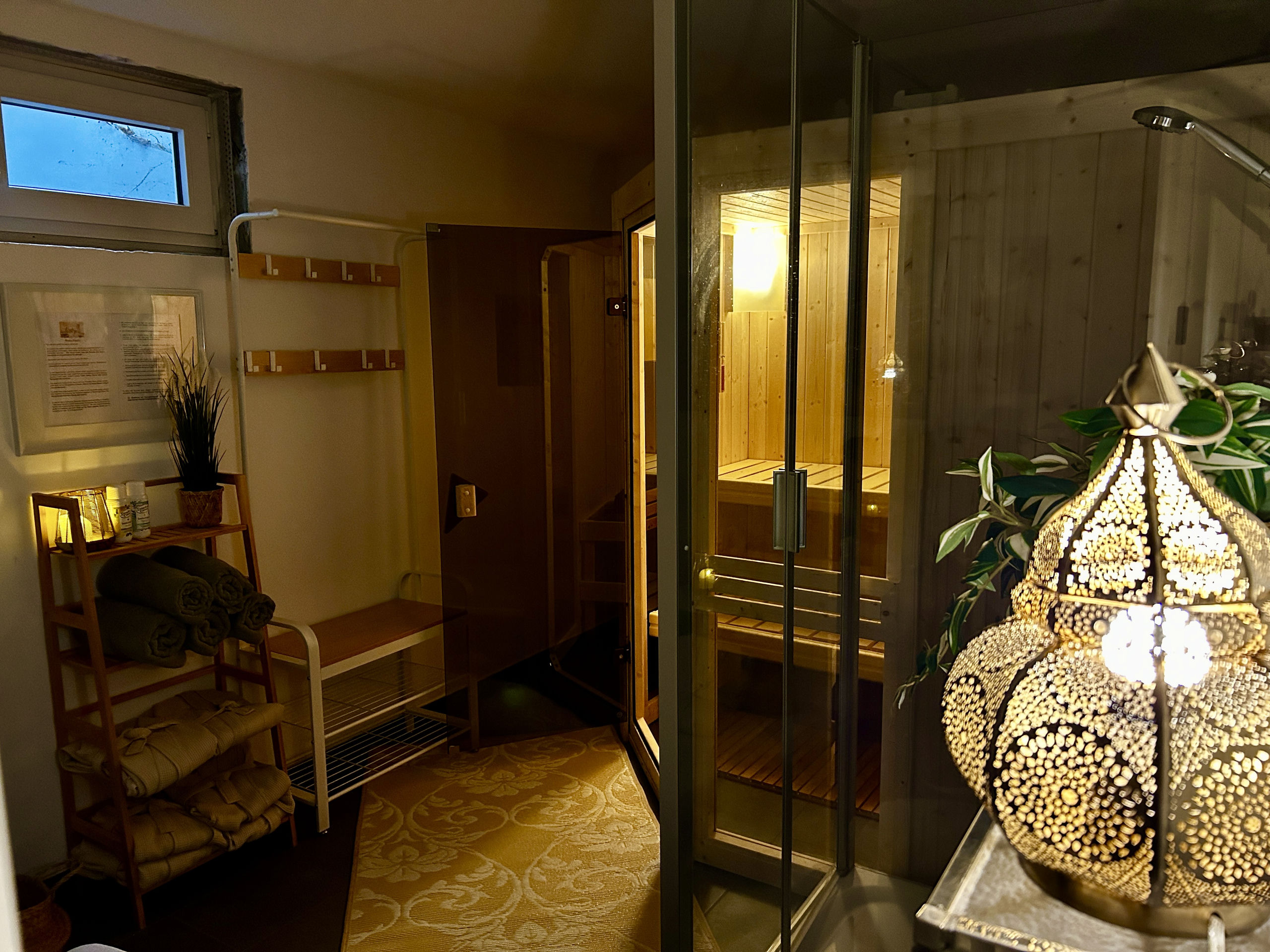 Der Spabereich mit beleuchteter Glastürsauna, einem Regal mit gerollten Handtüchern und gefalteten Bademänteln, Holzhaken an der Wand, Pflanzen und einer leuchtenden Messinglampe.