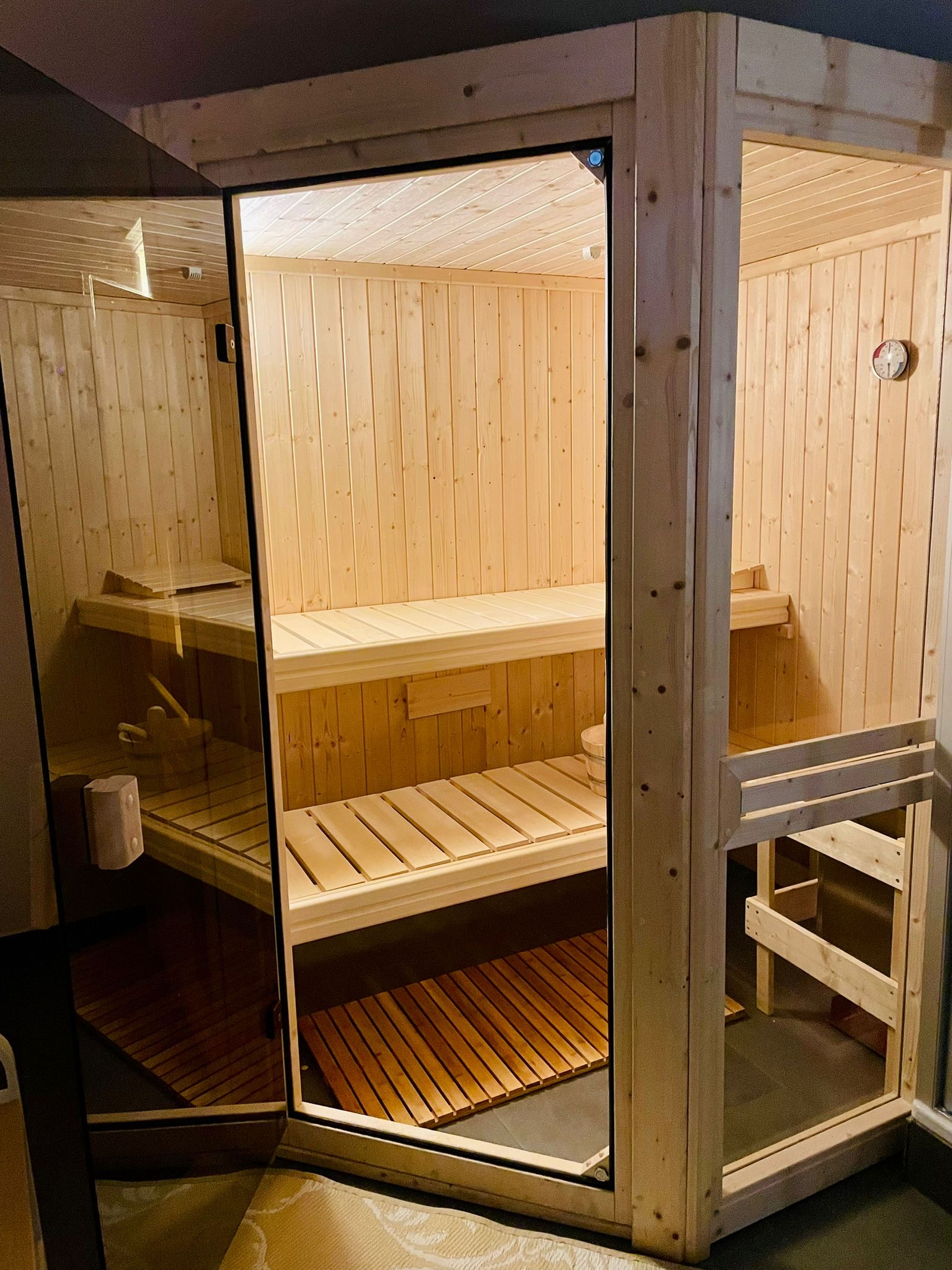 Saunakabine aus hellem Kiefernholz mit zwei Lattenbänken, einer Glastür mit Holzgriff, einem Holzeimer und rundem Thermometer an der Wand.