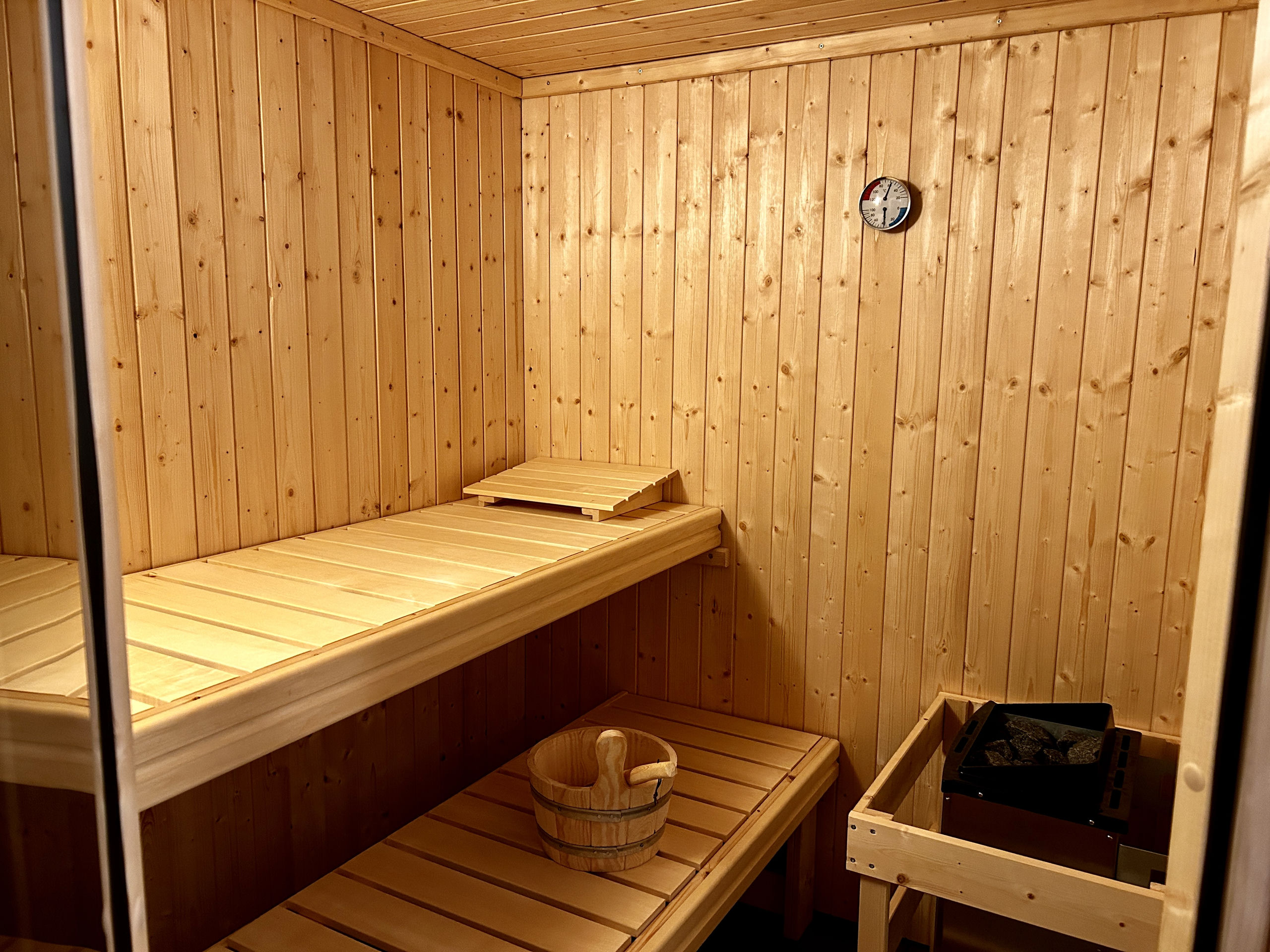 Innenansicht der Sauna mit hellen Kiefernholzwänden, zwei Lattenbänken mit kleinem Aufsatz, einem Holzeimer mit Kelle und dem Ofen hinter der Holzumrandung.