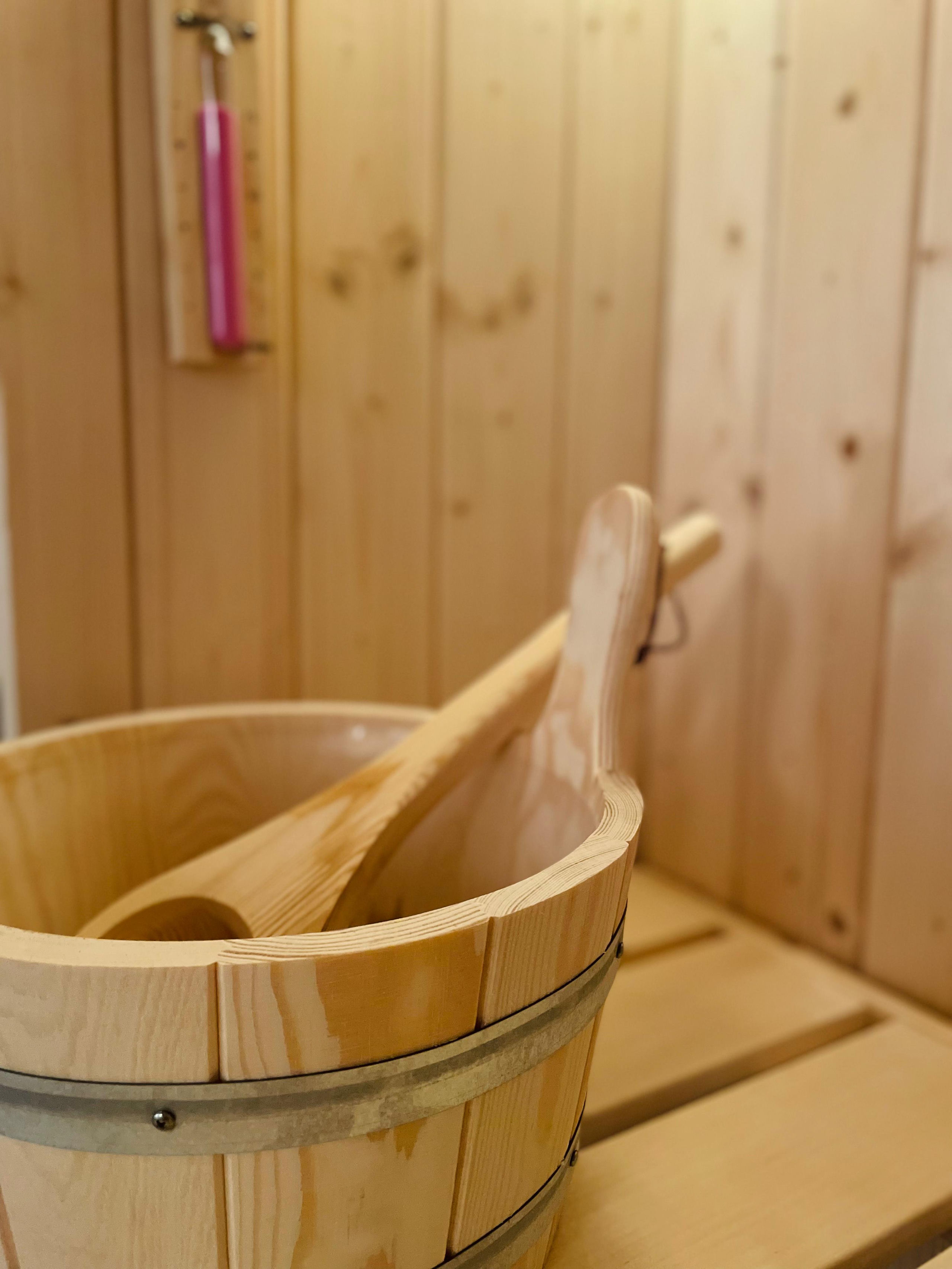 Nahaufnahme eines leeren Holzeimers mit Kelle auf der Lattenbank der Sauna, dahinter unscharf eine Sanduhr an der Holzwand.
