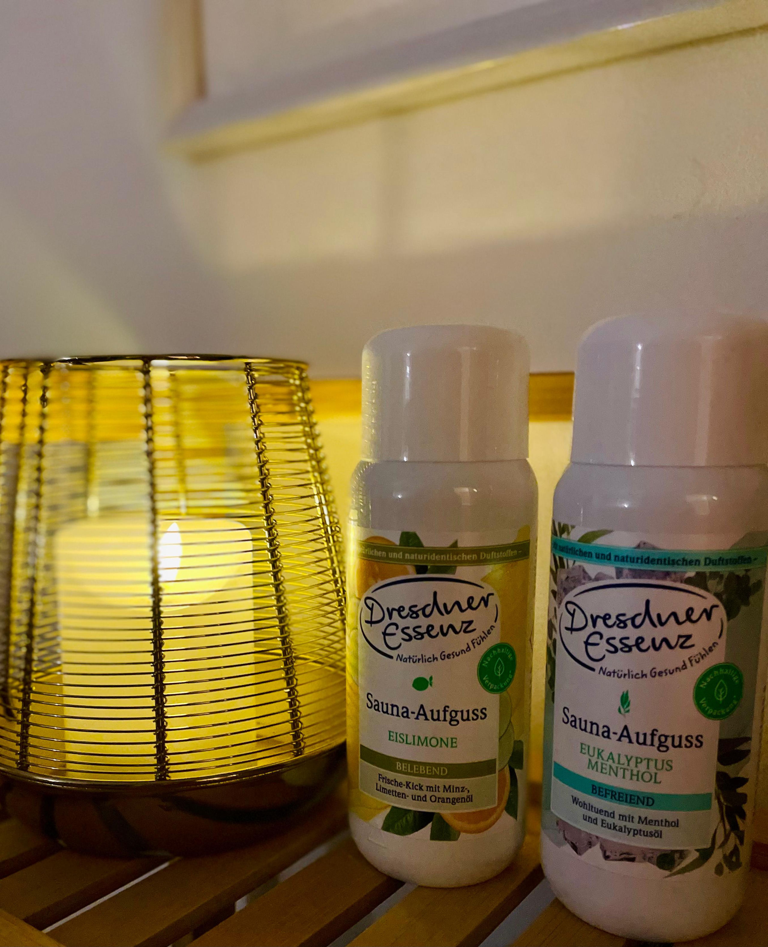 Zwei Flaschen Dresdner Essenz Sauna-Aufguss, Eislimone und Eukalyptus Menthol, stehen auf einem Lattenregal aus Bambus neben einer Kerze im goldenen Drahthalter.