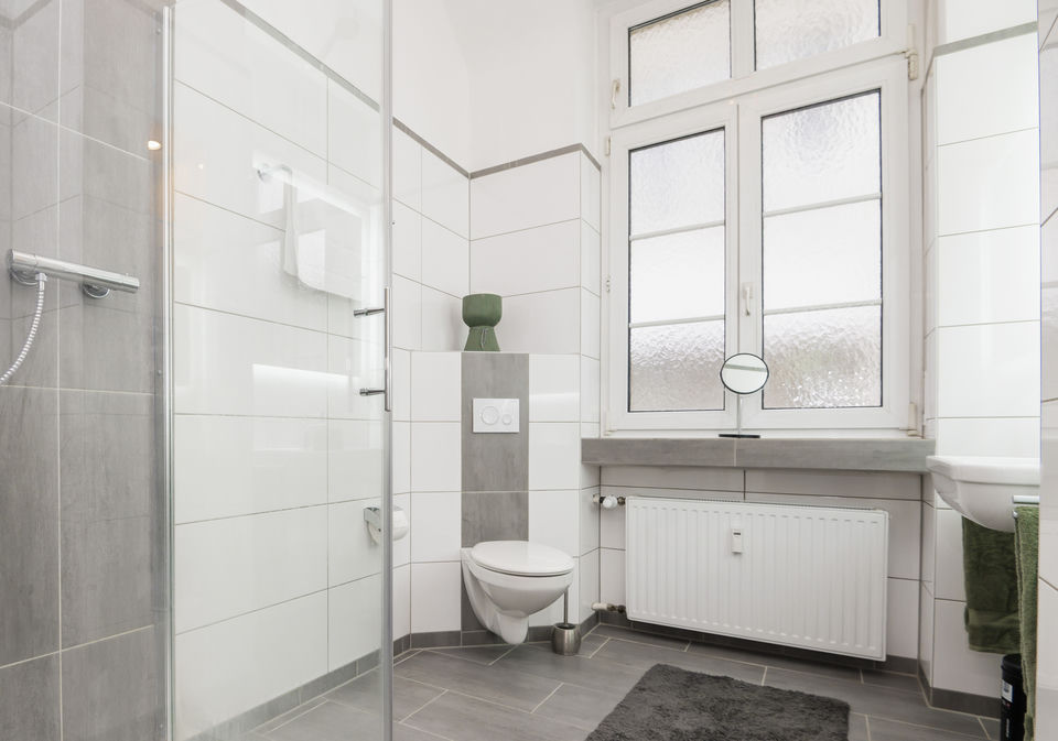 Helles Badezimmer mit Glasduschwand, wandhängendem WC, Milchglasfenster und weißem Heizkörper unter der grauen Steinablage.