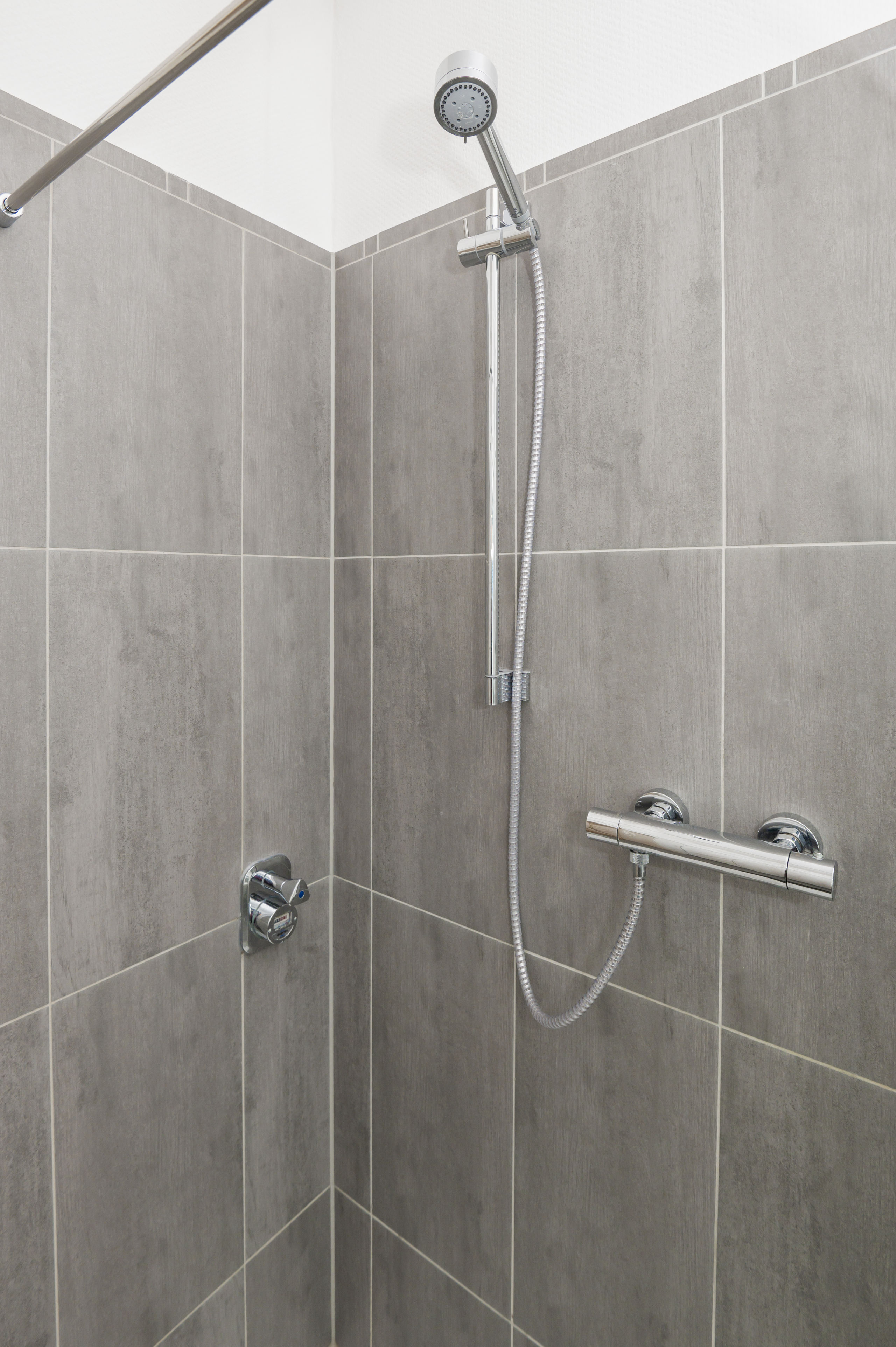 Detailaufnahme der ebenerdigen Dusche mit großformatigen grauen Fliesen, Chrom-Handbrause am Brausestab und sichtbarer Armatur.