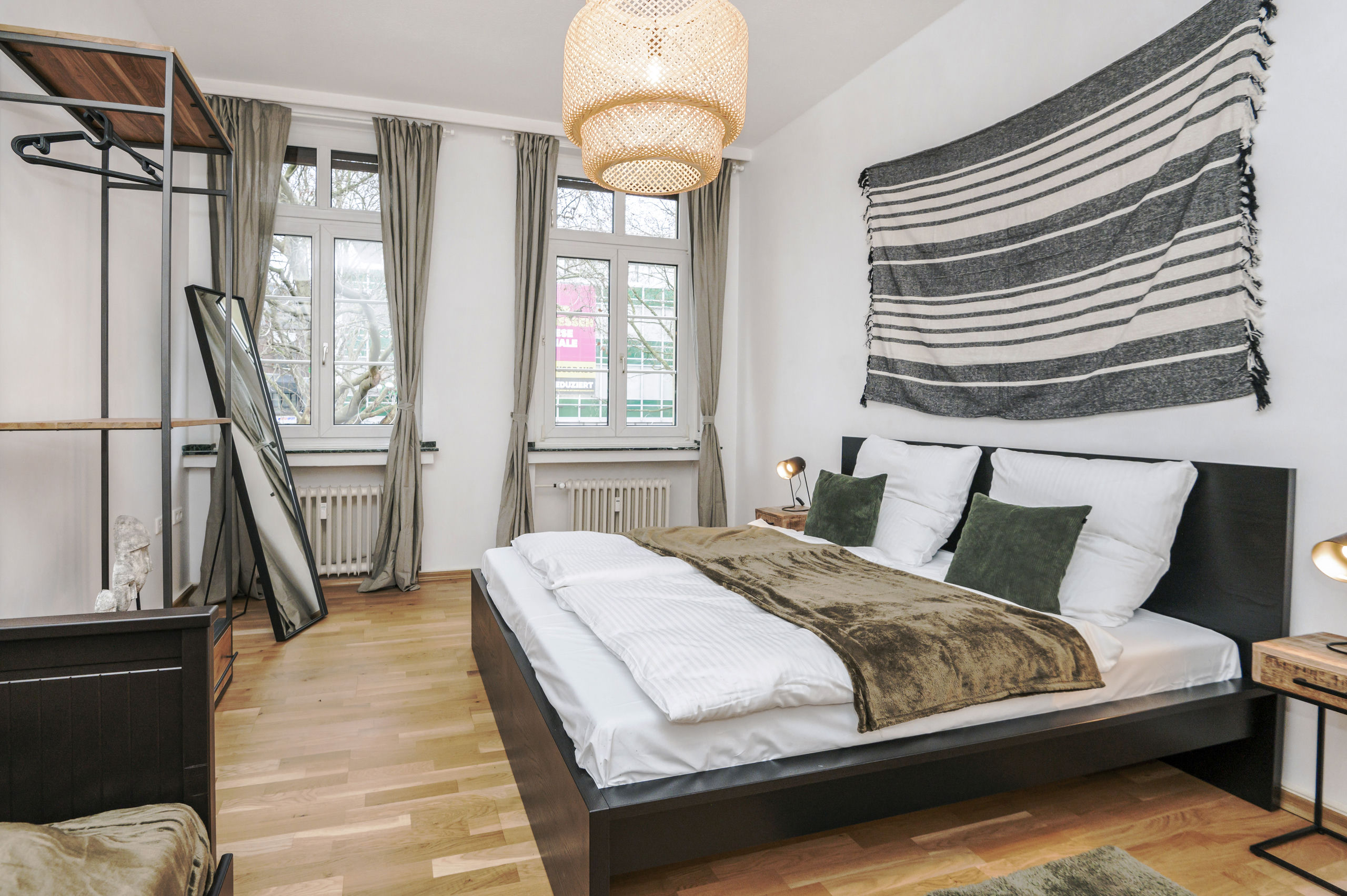 Doppelbett mit dunklem Holzrahmen in einem hellen Schlafzimmer, dazu hohe Fenster, ein offenes Kleidergestell und ein gestreifter Wandteppich.