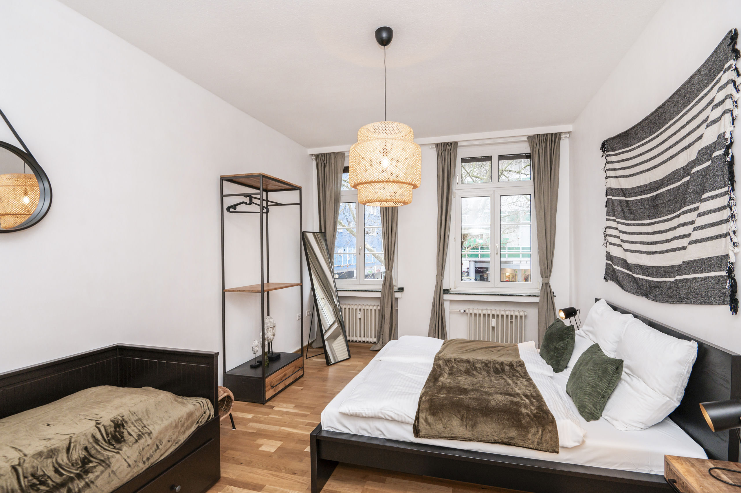 Blick von der anderen Seite: Doppelbett, Singlebett mit Bettkasten, offenes Kleidergestell, Anlehnpiegel und eine Rattan-Hängeleuchte über dem Parkett.