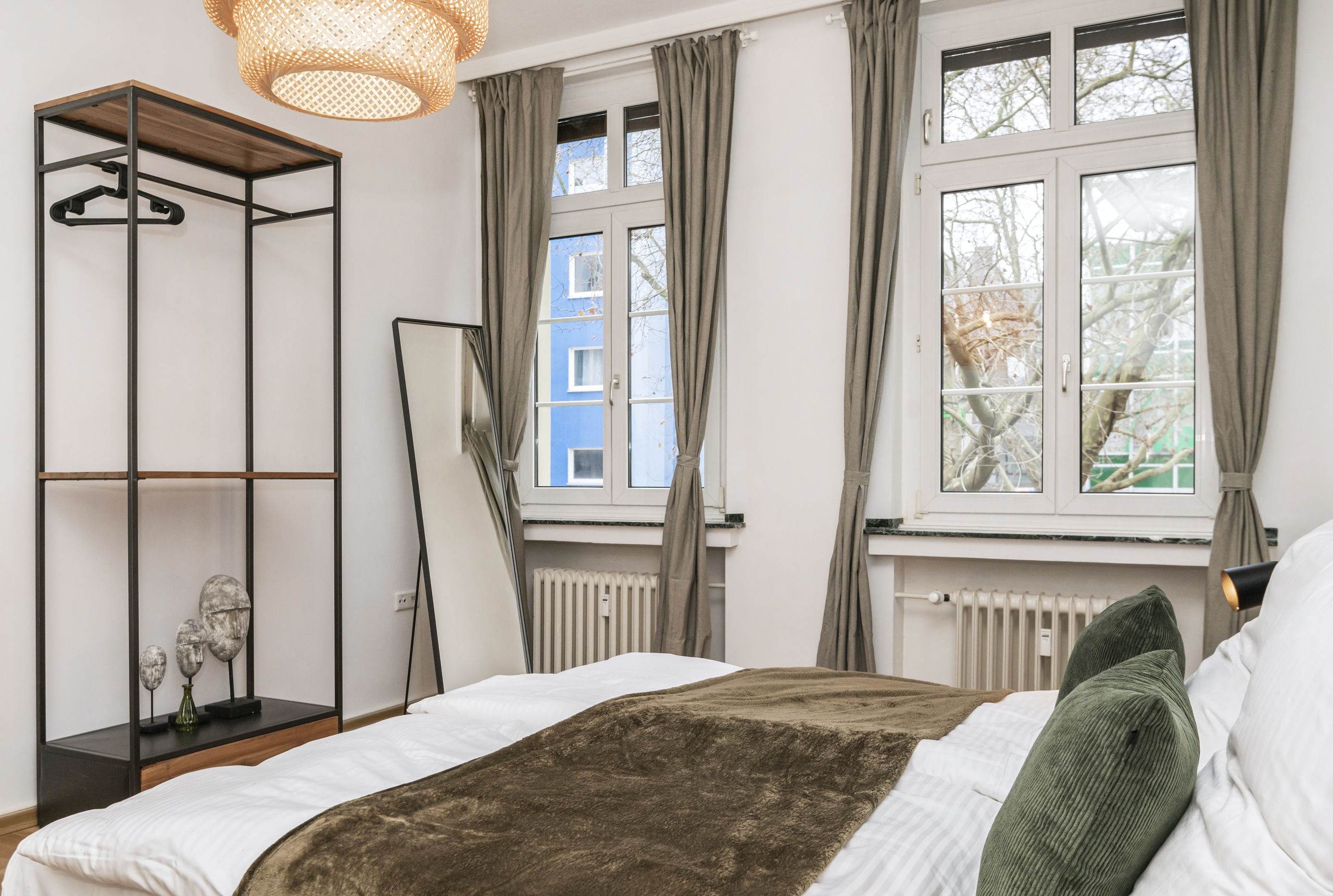 Bettfußende mit Blick zu zwei hohen Fenstern mit graubeigen Gardinen, daneben ein offenes Kleiderregal und ein hoher Anlehnpiegel.