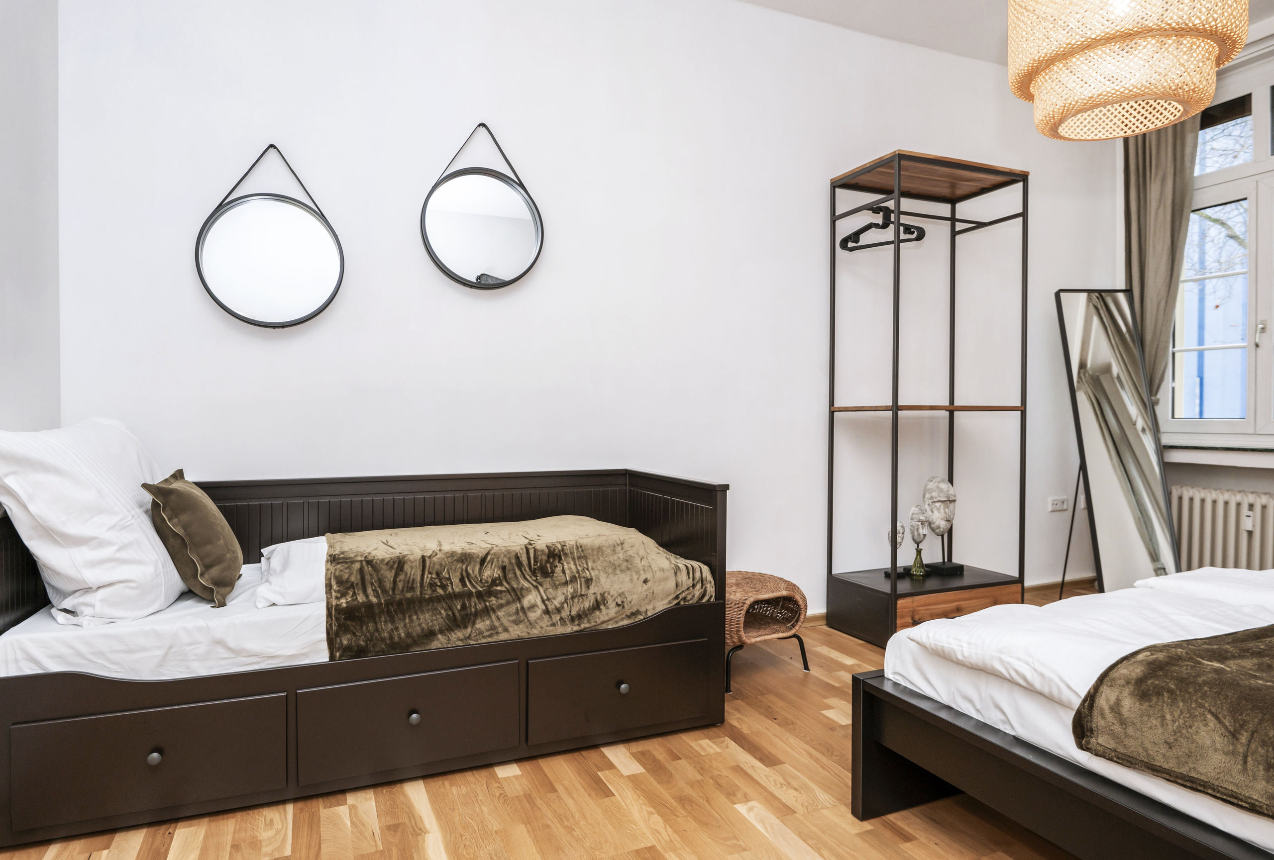 Singlebett mit Auszugsschubladen und zwei runden Hängespiegeln darüber an der weißen Wand, daneben das offene Kleidergestell und ein Anlehnpiegel auf hellem Parkett.