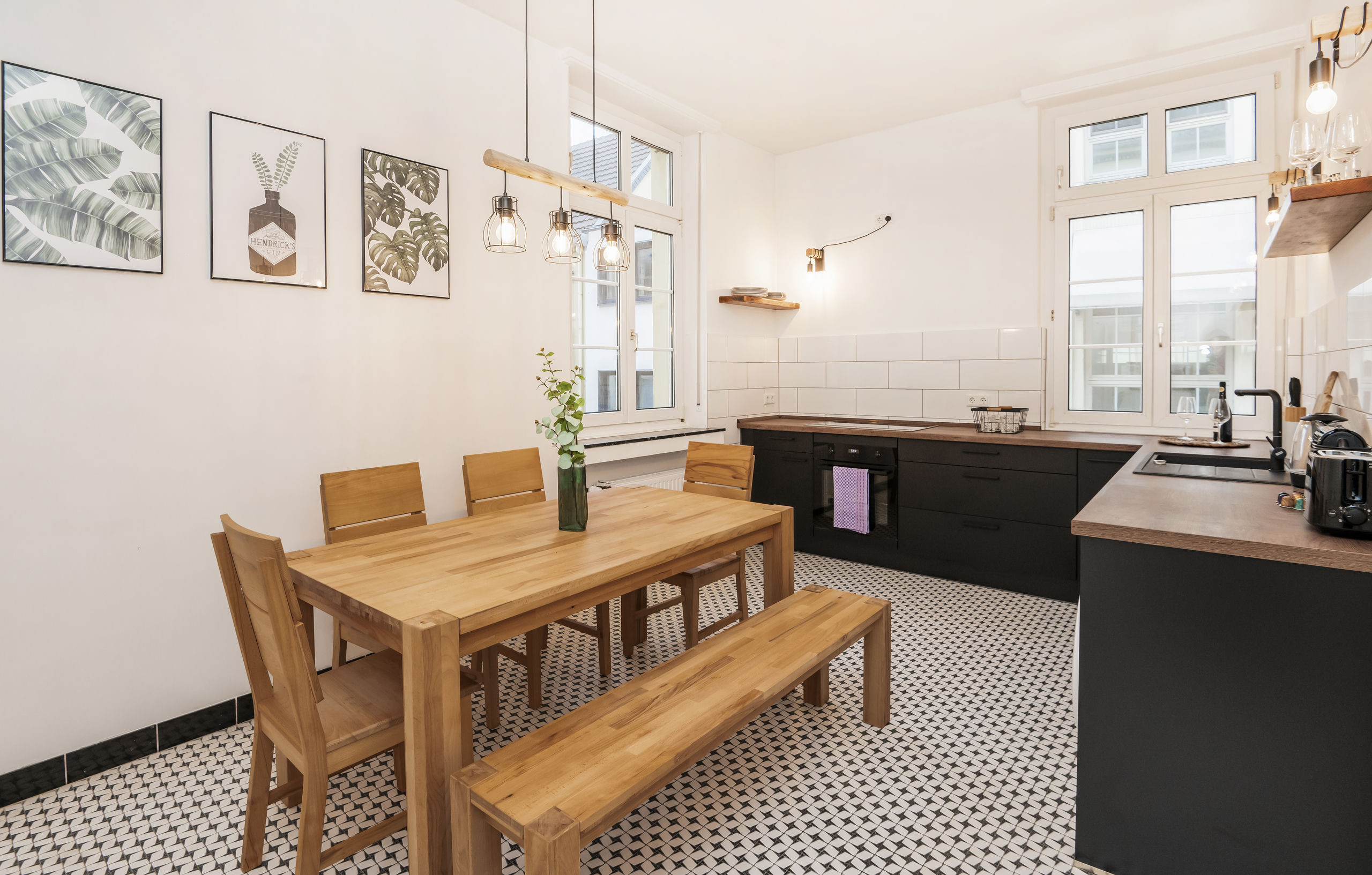Cocina con mesa de comedor de madera maciza y banco, muebles bajos negros, encimeras de madera y suelo de baldosas en blanco y negro.
