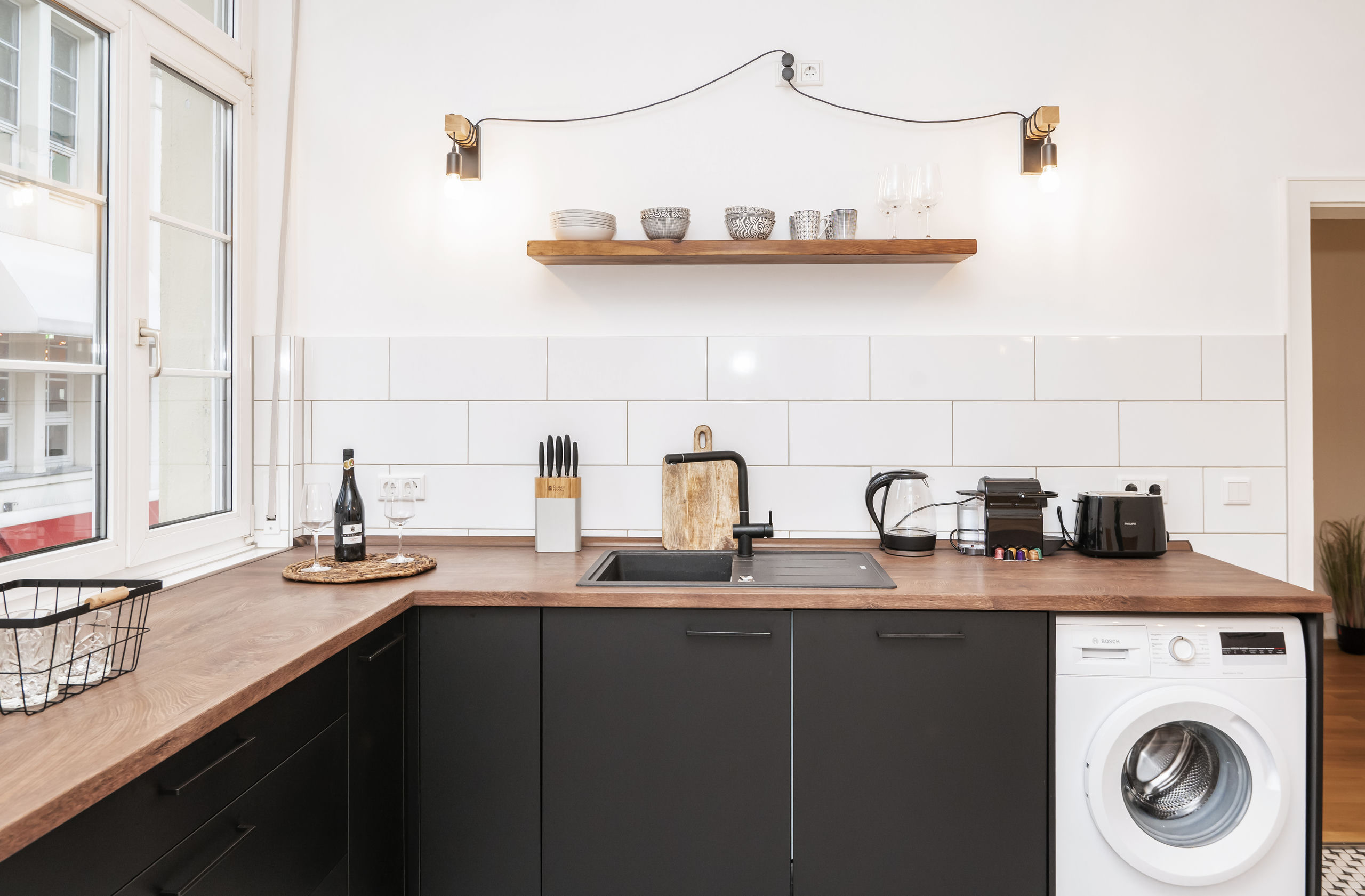 Küchenecke mit mattschwarzen Schränken und Holz-Arbeitsplatte, schwarzem Spülbecken, offenem Holzregal, Wasserkocher, Kaffeemaschine und Waschmaschinenplatz.