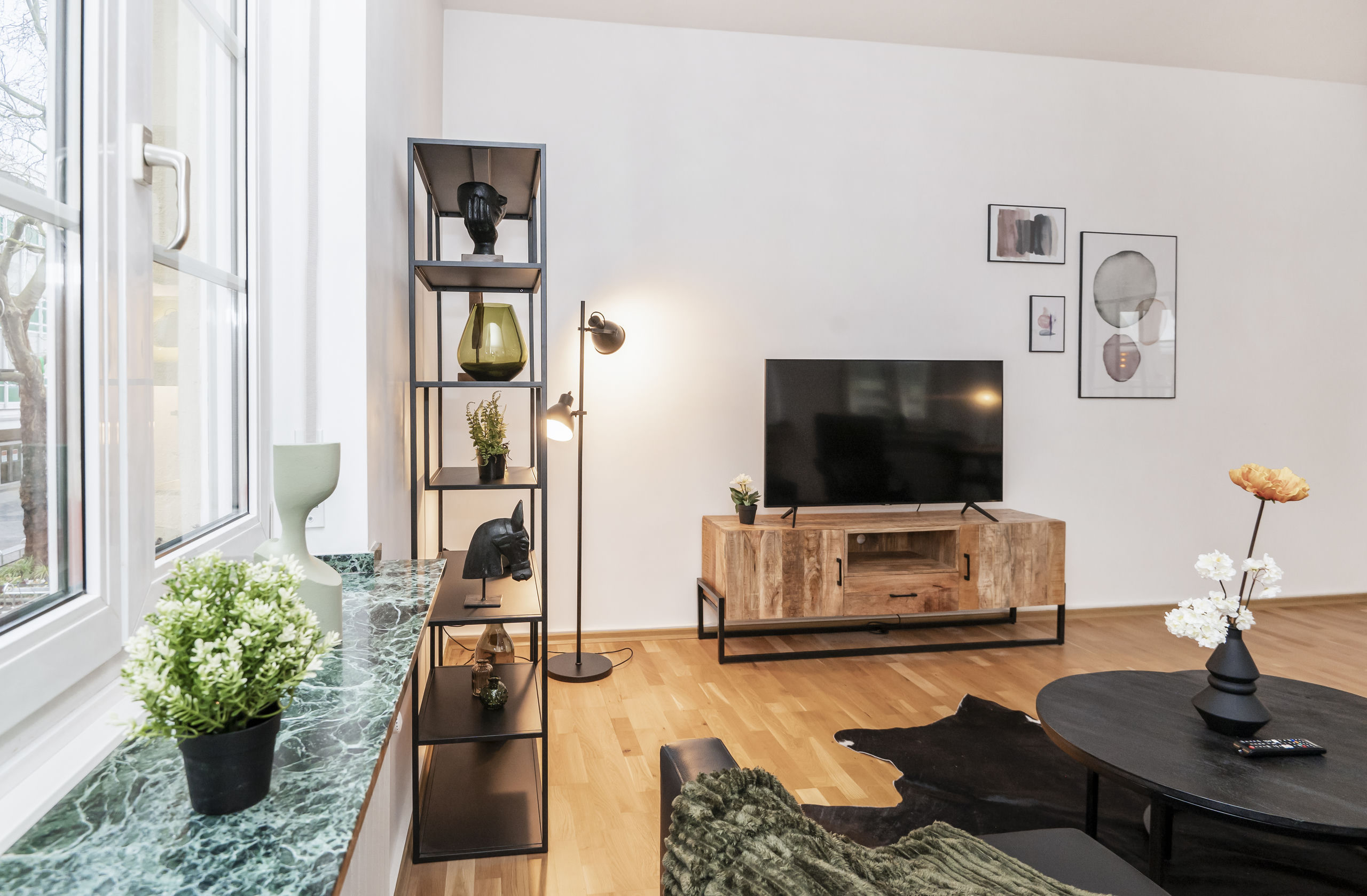 Wohnzimmer mit Flachbildschirm auf Holzsideboard, hohem schwarzem Regal mit Dekoobjekten und grünem Marmorfensterbrett über Parkettboden.