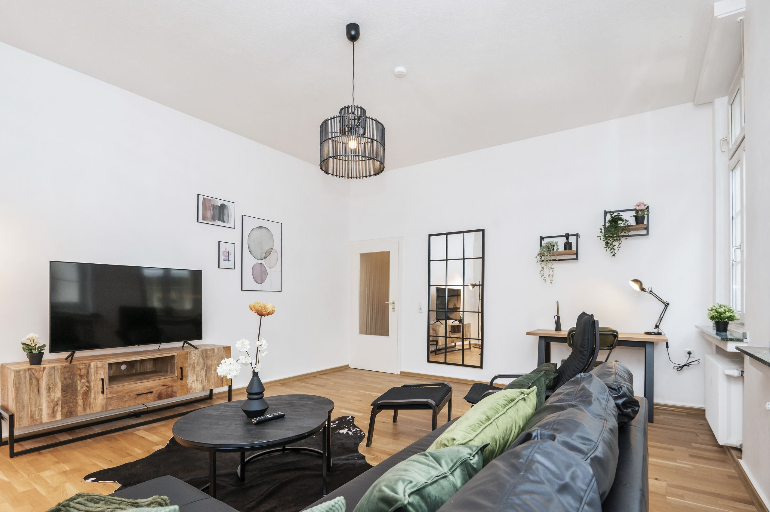 Graues Ledersofa mit grünen Kissen vor dem Fernseher, dazu schwarze Pendelleuchte, Anlehnpiegel und ein Schreibtisch am Fenster.