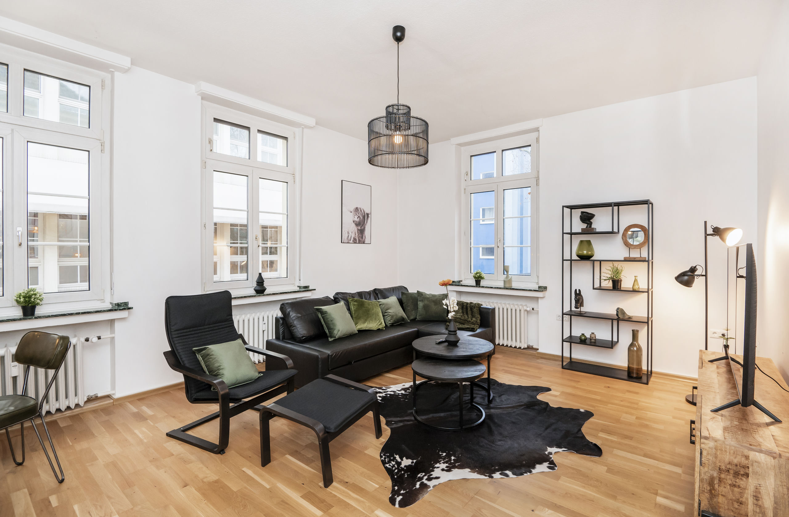 Sitzecke mit dunklem Ecksofa, schwarzem Sessel mit Hocker und runden Beistelltischen auf einem Fellteppich vor hohen Fenstern.