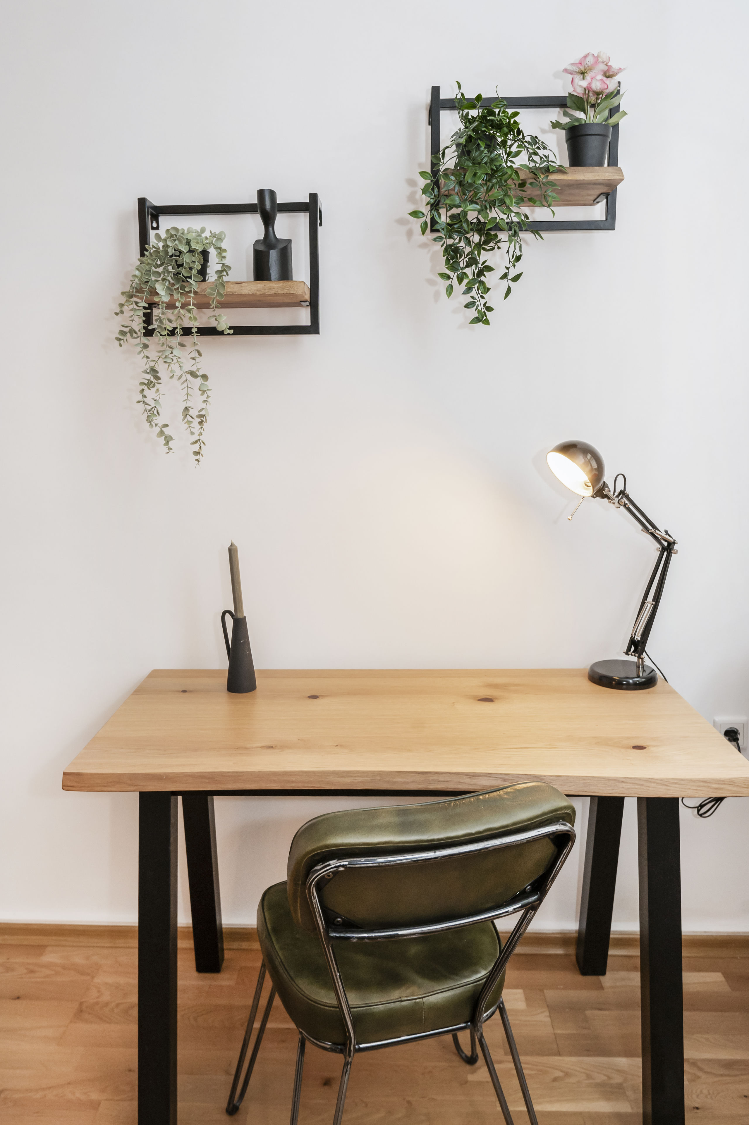 Schreibtisch aus hellem Holz mit schwarzem Gestell und olivgrünem Lederstuhl vor weißer Wand, darüber zwei Regalbretter mit Pflanzen.