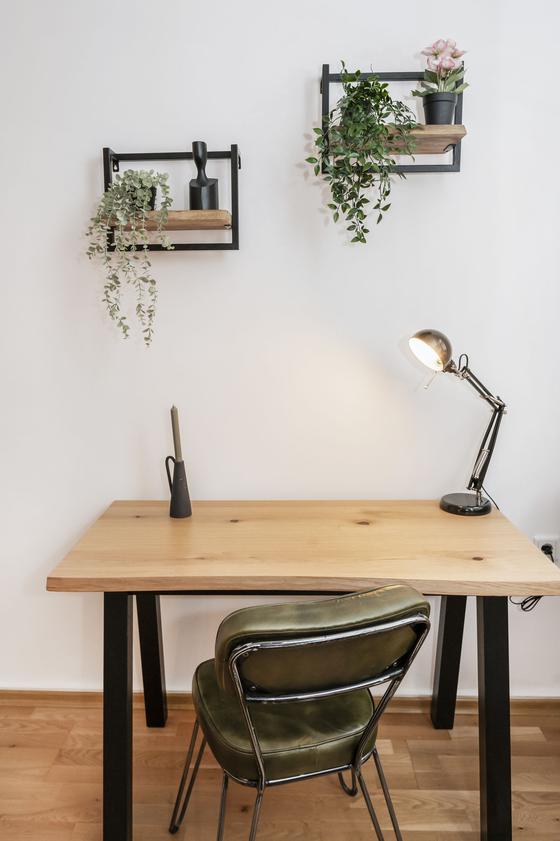 Wooden desk with black legs and an olive leather chair against a white wall, with a black task lamp and two plant shelves above.