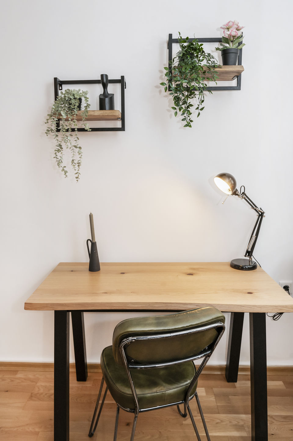 Schreibtisch aus hellem Holz mit schwarzem Gestell und olivgrünem Lederstuhl vor weißer Wand, darüber zwei Regalbretter mit Pflanzen.
