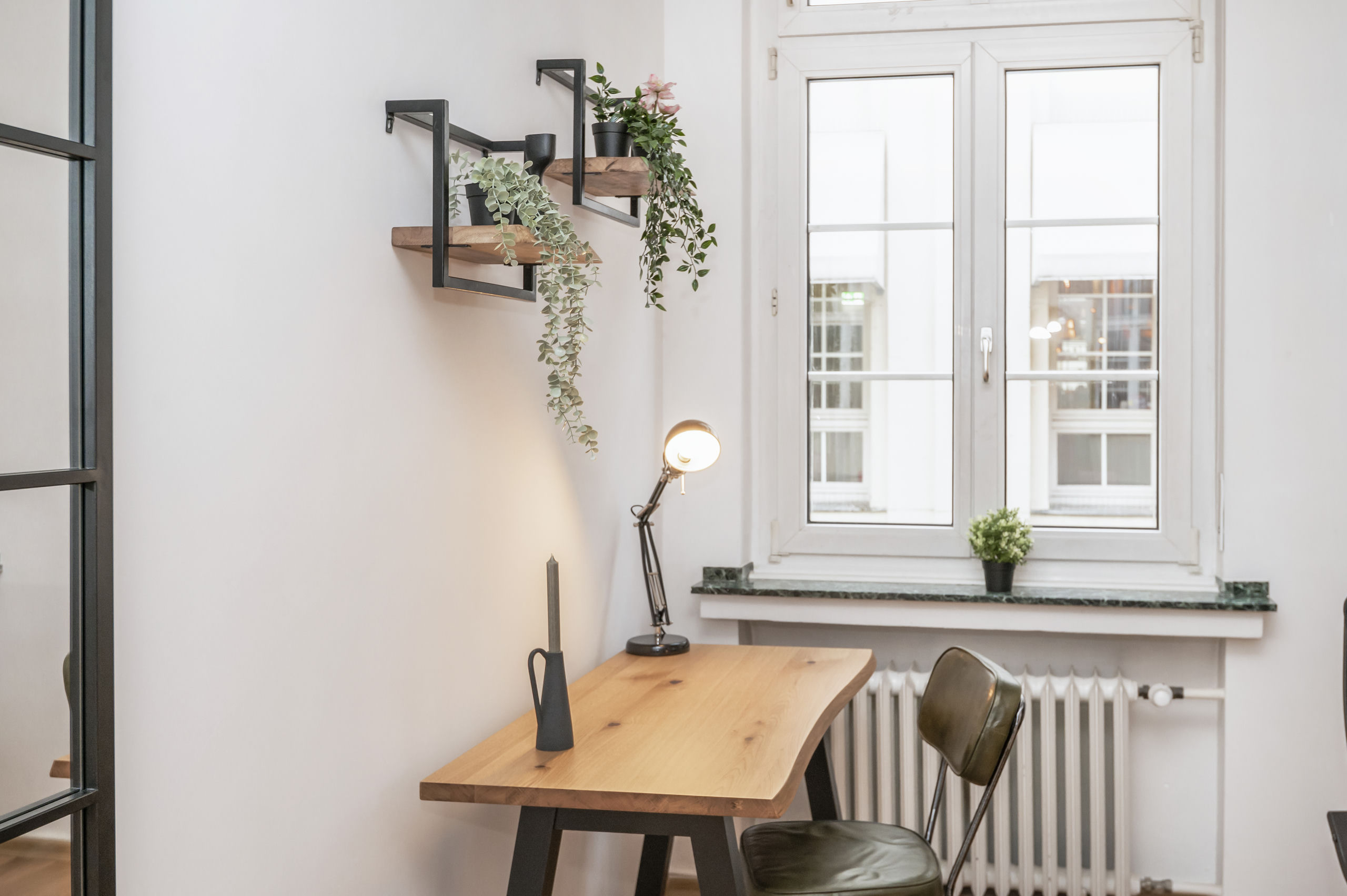 Seitenansicht des Schreibtischs am hohen Fenster mit Marmorfensterbank, darunter ein Heizkörper und Hängepflanzen an der Wand.