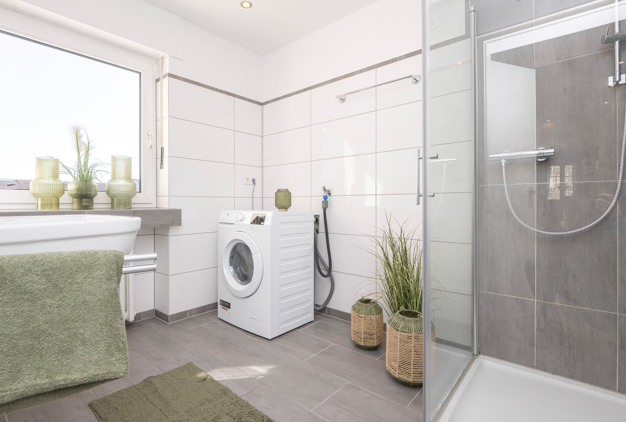 Bright bathroom with a glass-fronted walk-in shower, white tiled walls, a washbasin beneath the window and a washing machine in the corner.