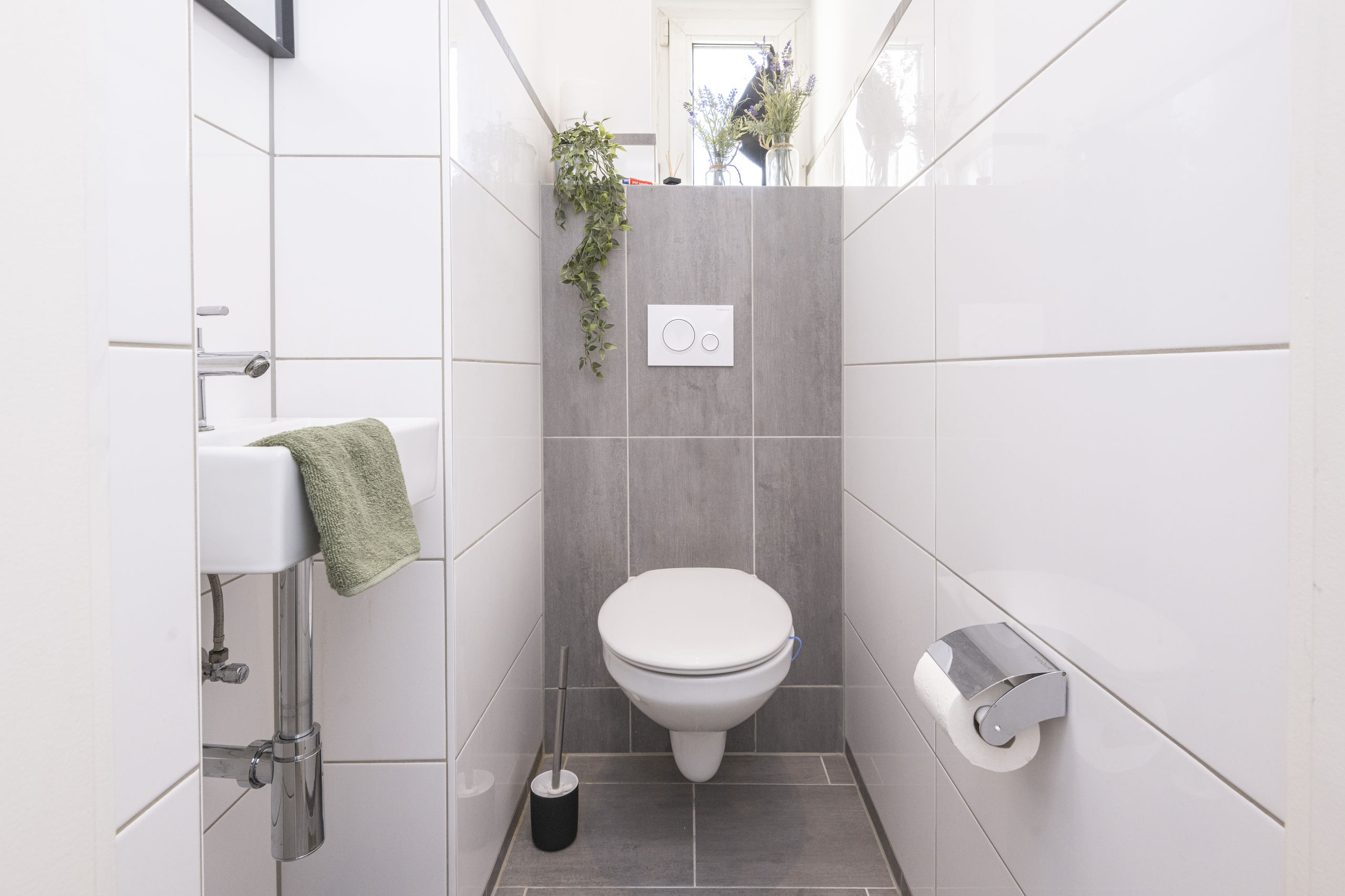Compact guest toilet with a wall-hung bowl in front of a grey tiled niche and a small white basin to the side.