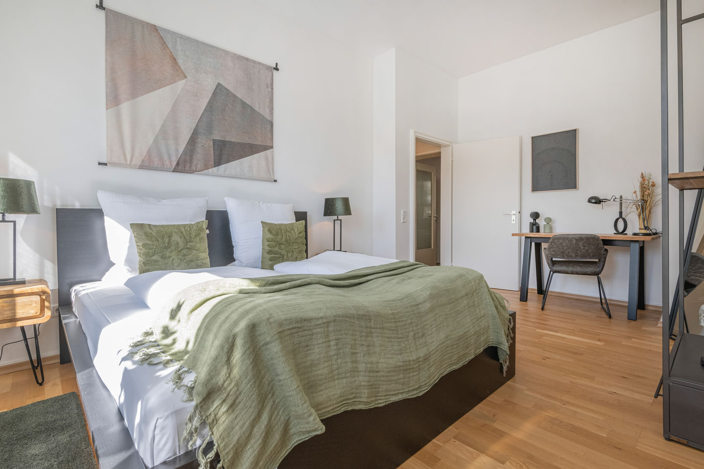 Dormitorio con cama de matrimonio con ropa de cama blanca, colcha verde salvia y cojines verdes, tapiz geométrico sobre el cabecero y escritorio junto a la puerta.