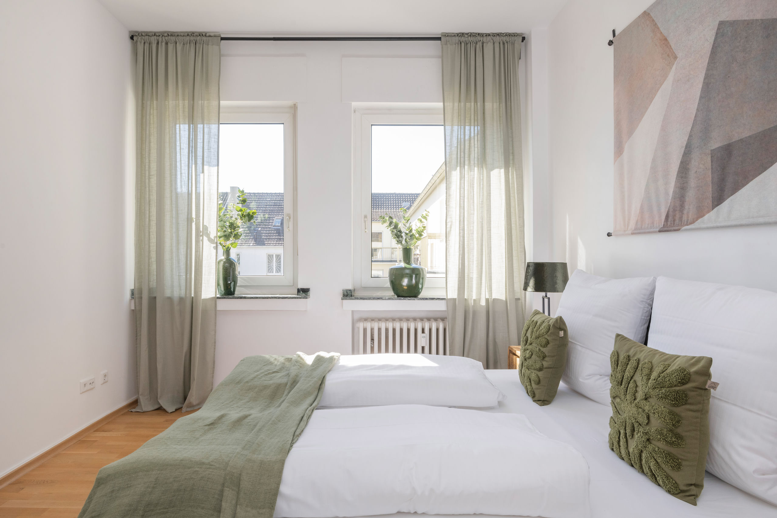 View across the made bed towards two tall windows with sheer curtains and green glass vases on the sills.