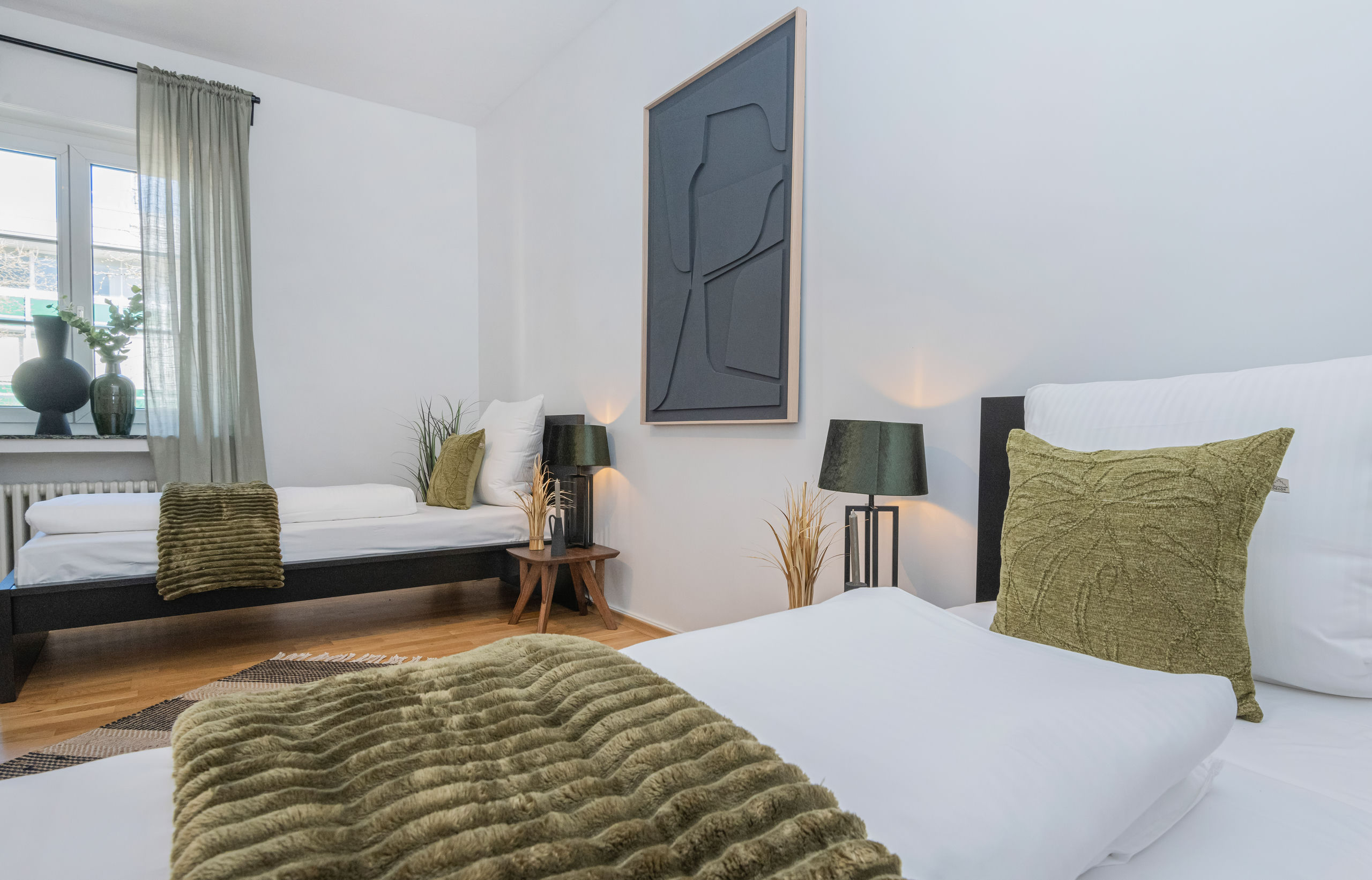 Bedroom with a double bed in the foreground and a low single bed by the window, both dressed in white with green cushions.