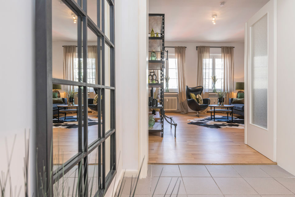 Hallway view through a black steel-framed glass partition into the living room with oak floors and a leather lounge chair.