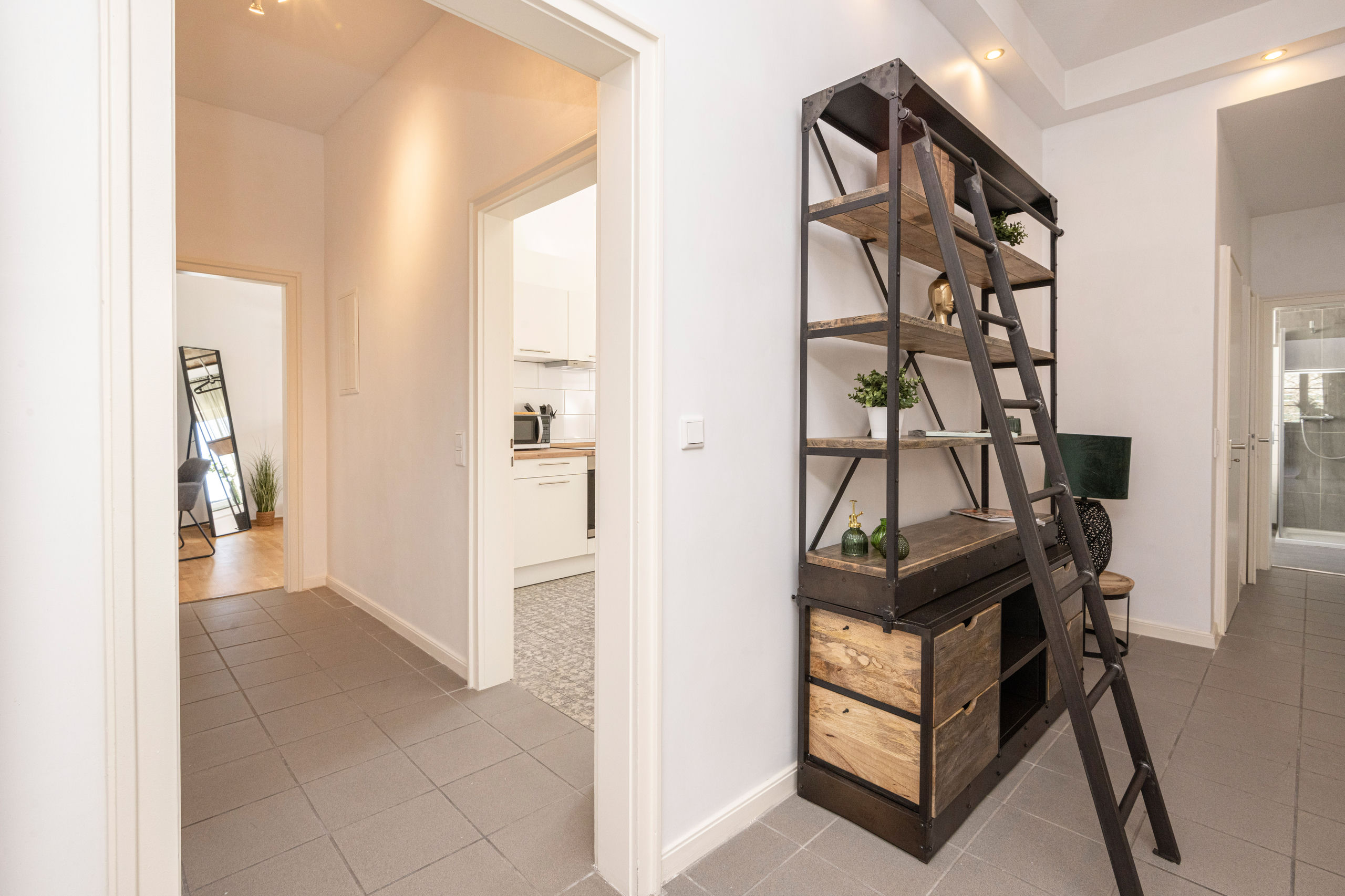 Tiled hallway with a tall industrial shelf and rolling ladder, doorways leading towards the kitchen and the bathroom.