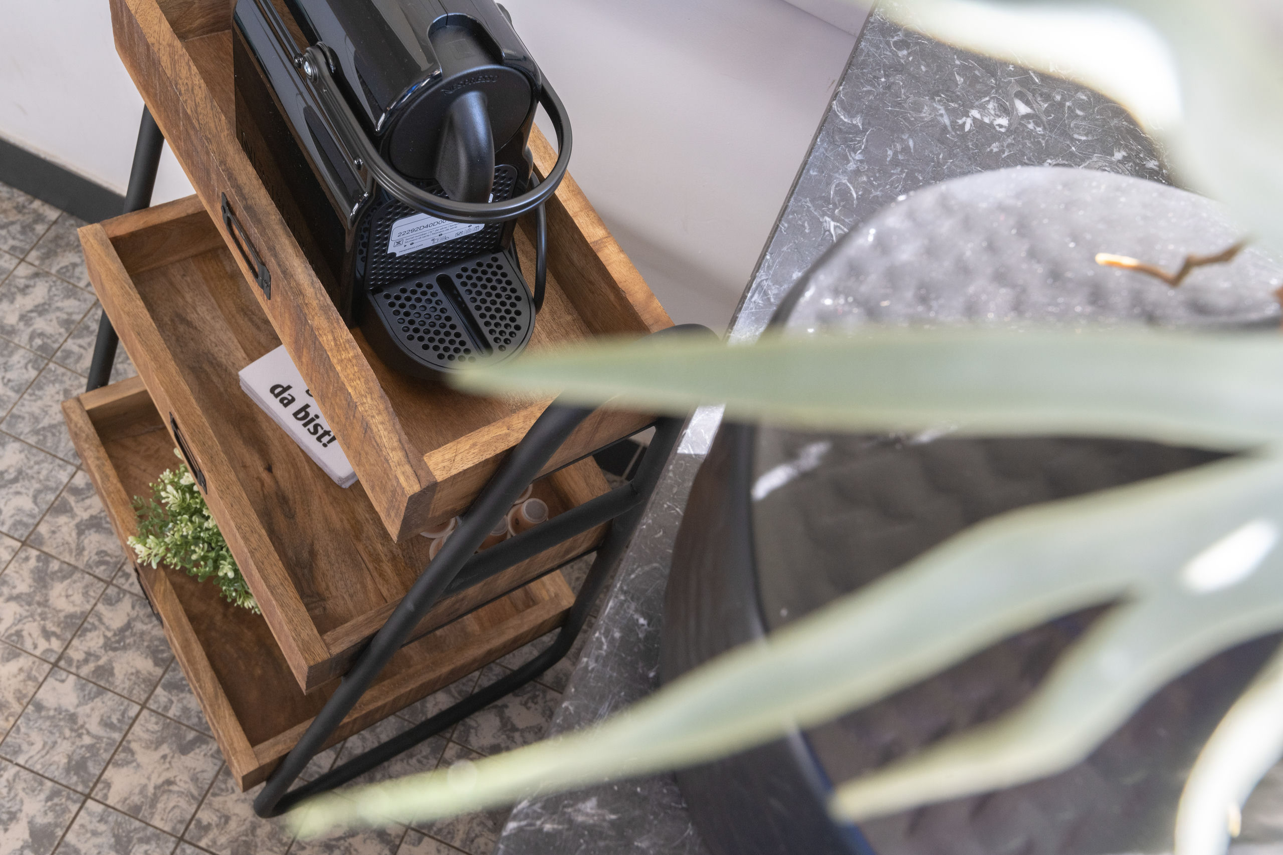 Close-up of a black capsule coffee machine on a wooden drawer trolley, with capsules and a small plant on the shelf below.