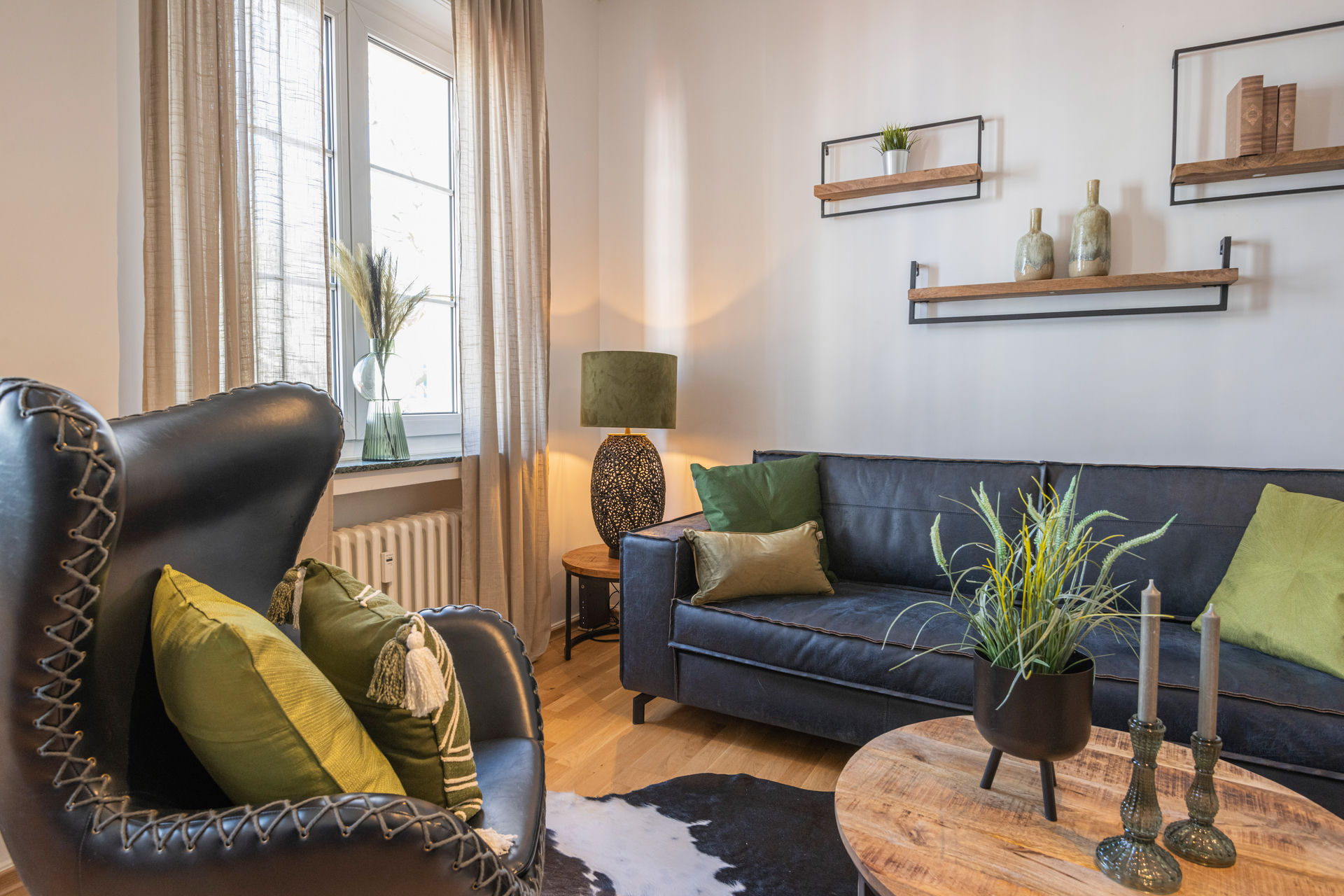 Living room with a dark leather wing chair, grey sofa with green cushions, wooden coffee table and wall-mounted open shelves.