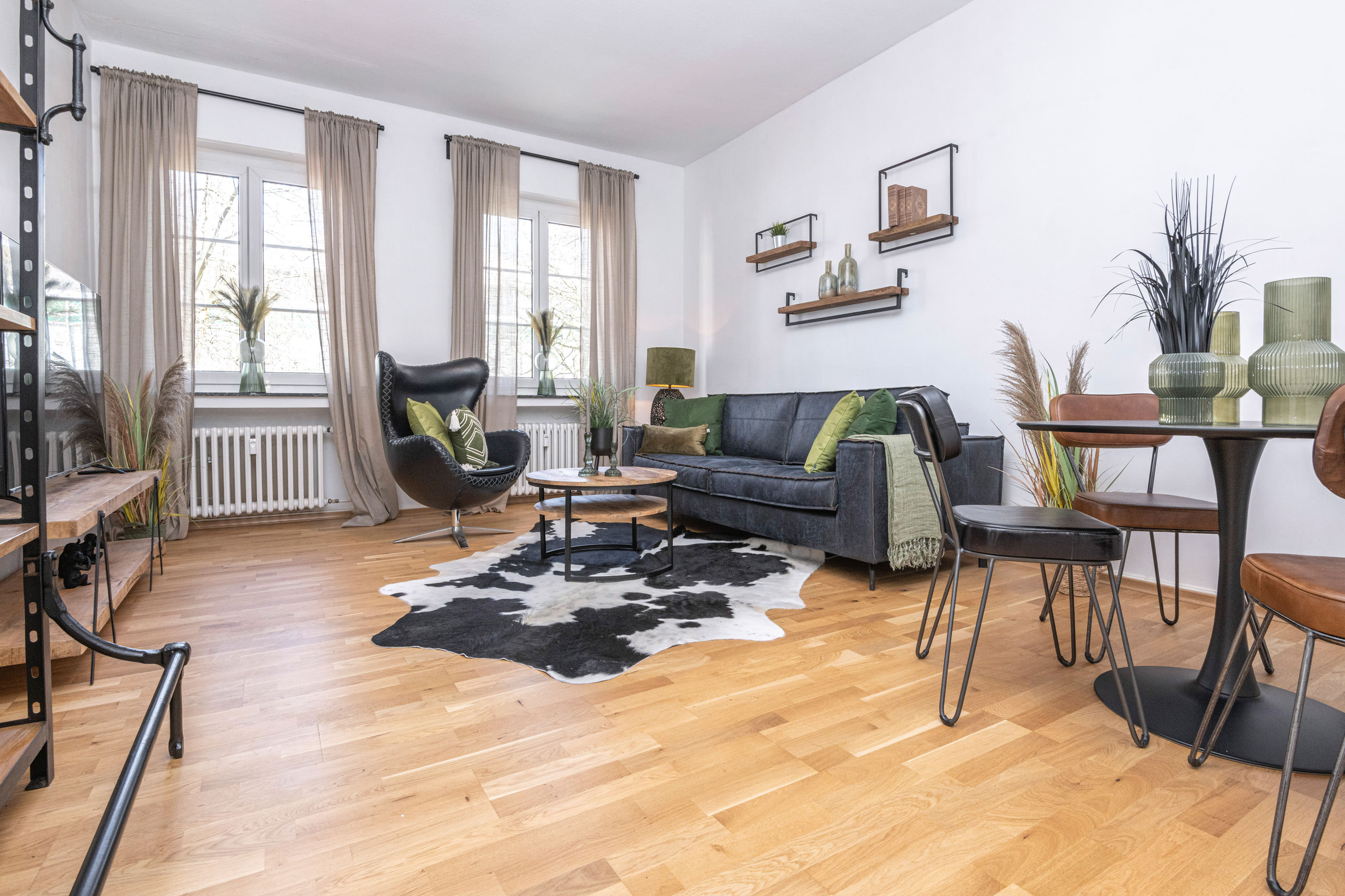 Wide view of the living room with two tall windows, a swivel leather chair, sofa and cowhide rug, plus a dining table.
