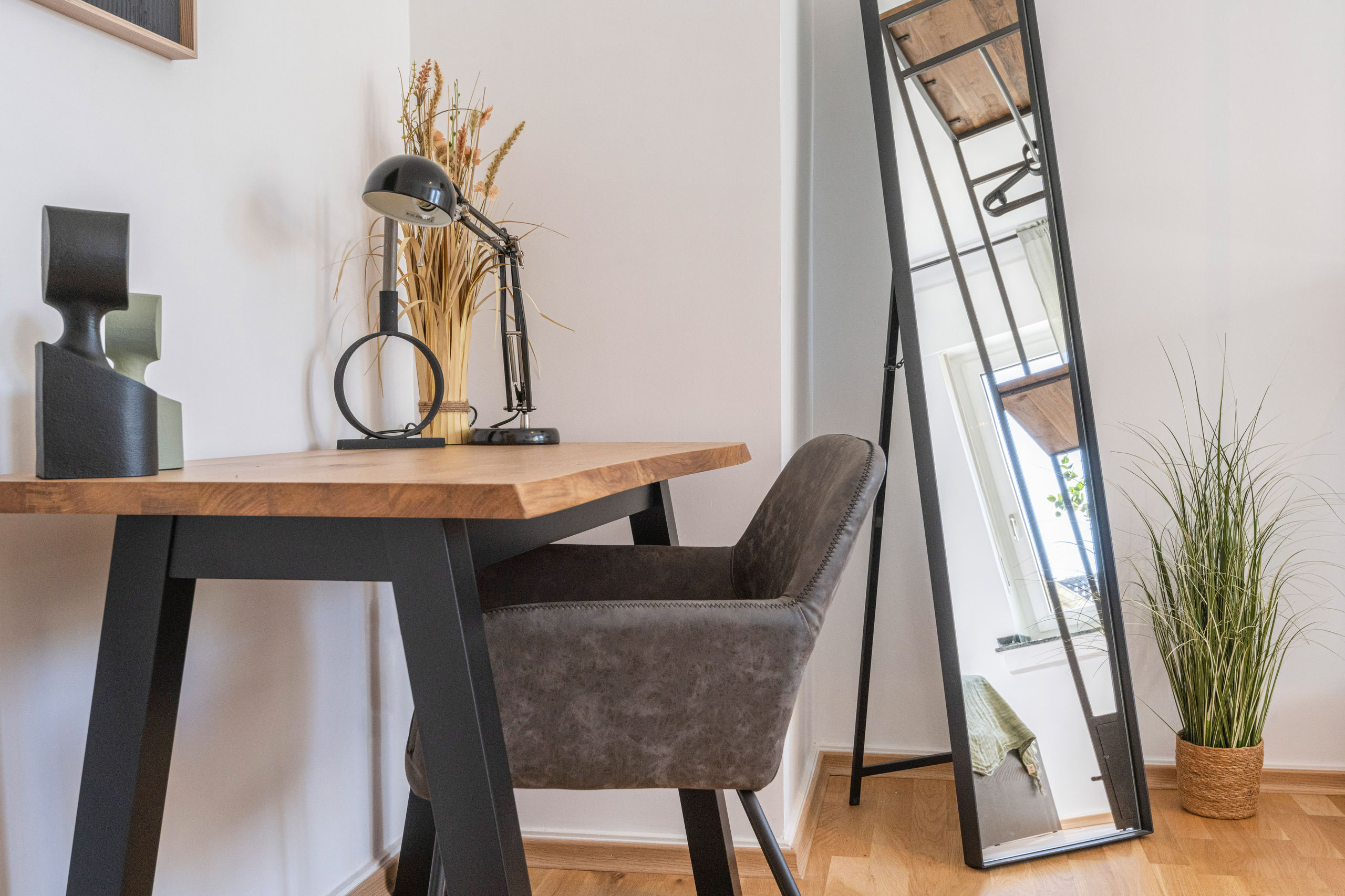 Home office corner with a wood-top desk, upholstered armchair, black adjustable lamp and a full-length mirror leaning against the wall.