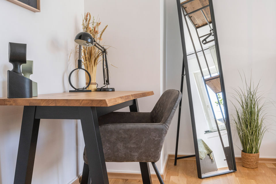 Home office corner with a wood-top desk, upholstered armchair, black adjustable lamp and a full-length mirror leaning against the wall.