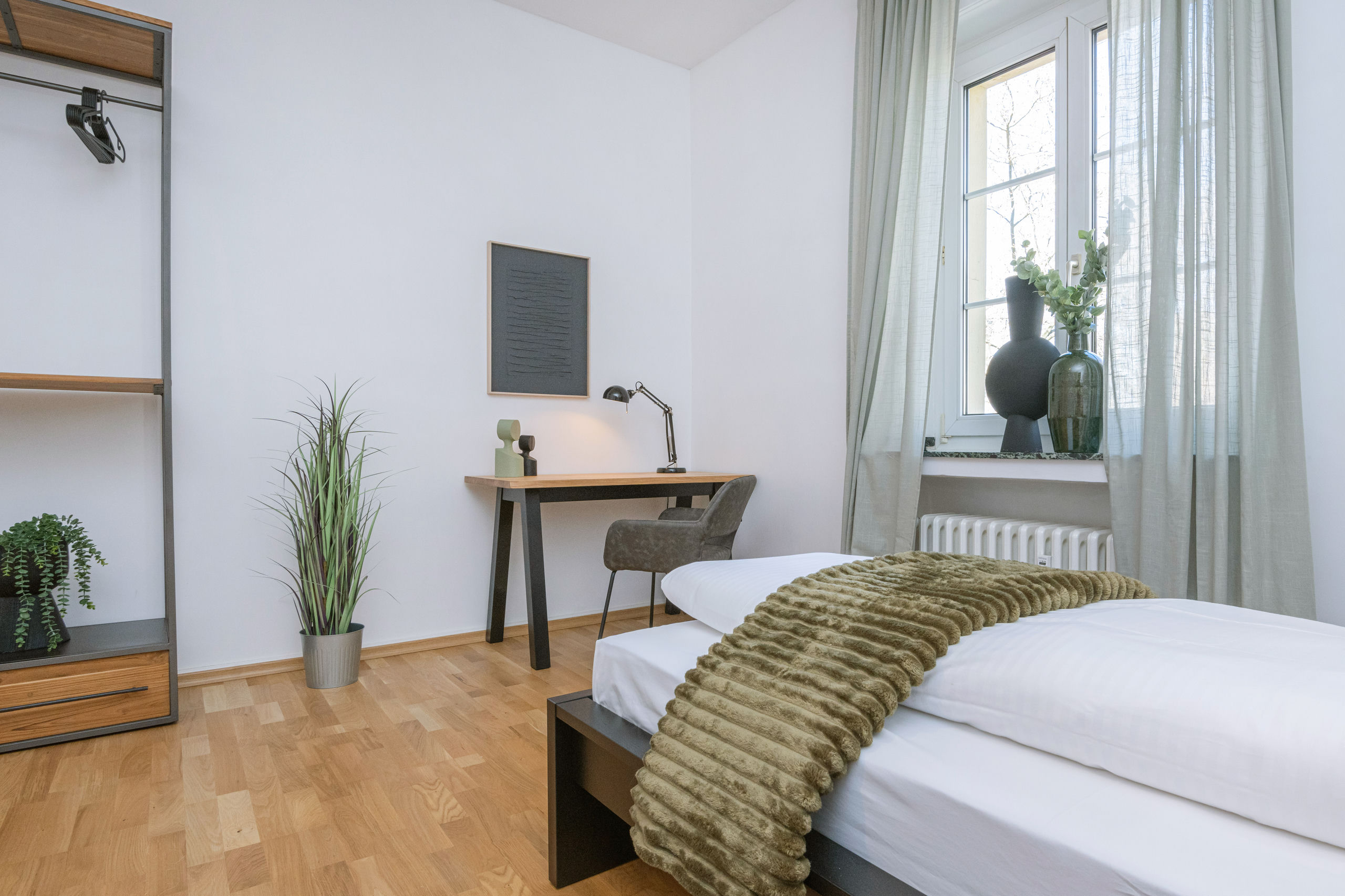 Desk and upholstered chair against a white wall in the bedroom, with the bed in the foreground and an open wardrobe beside it.