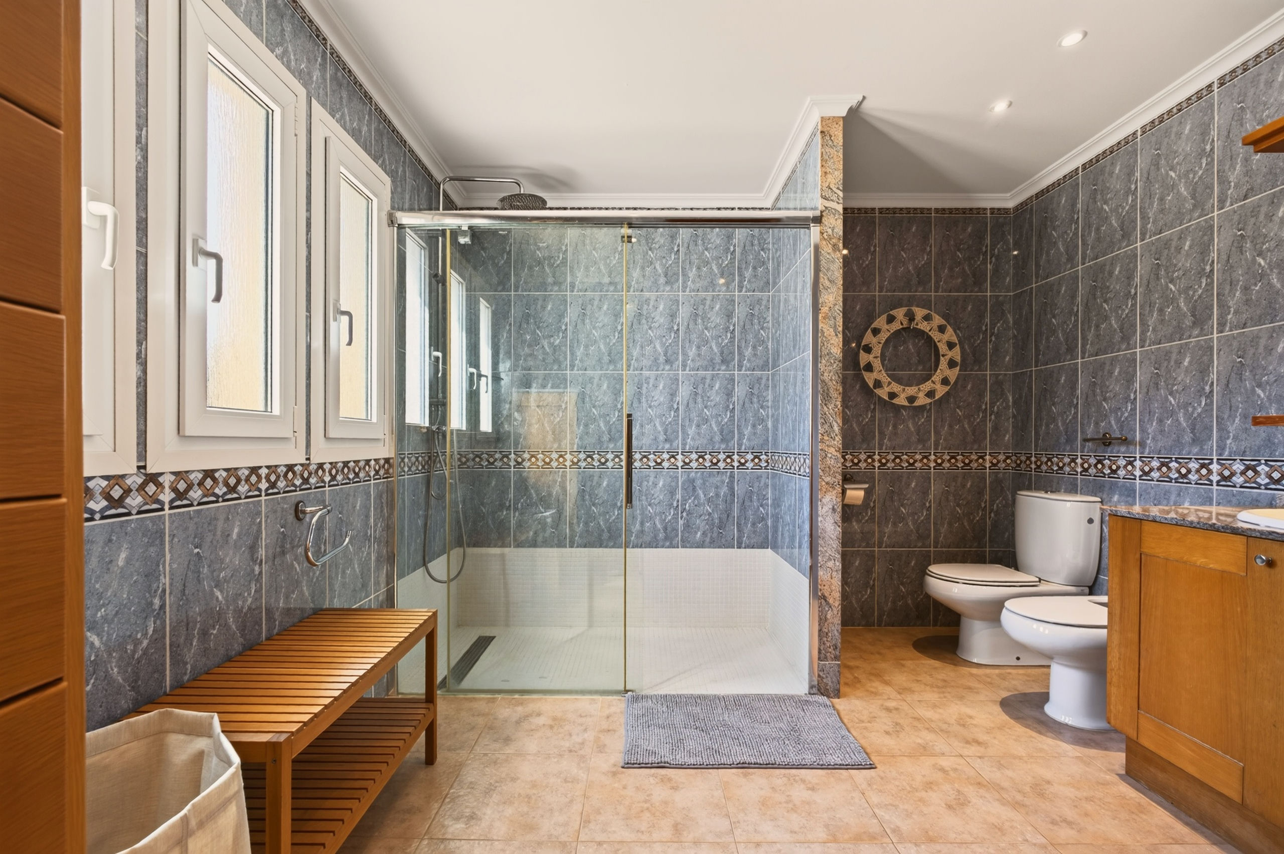 Großzügiges Bad mit grau marmorierten Fliesen, begehbarer Glas-Dusche mit Regenbrause, Holzbank, Toilette und Holzwaschbecken