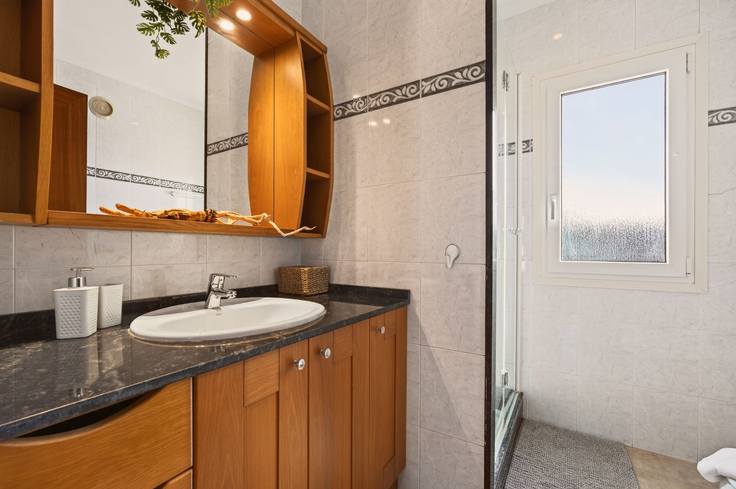 Nahaufnahme des Waschplatzes im hellen Bad: ovales Becken auf Granitplatte, Holzschubladen und Spiegelschrank, rechts die Duschkabine.