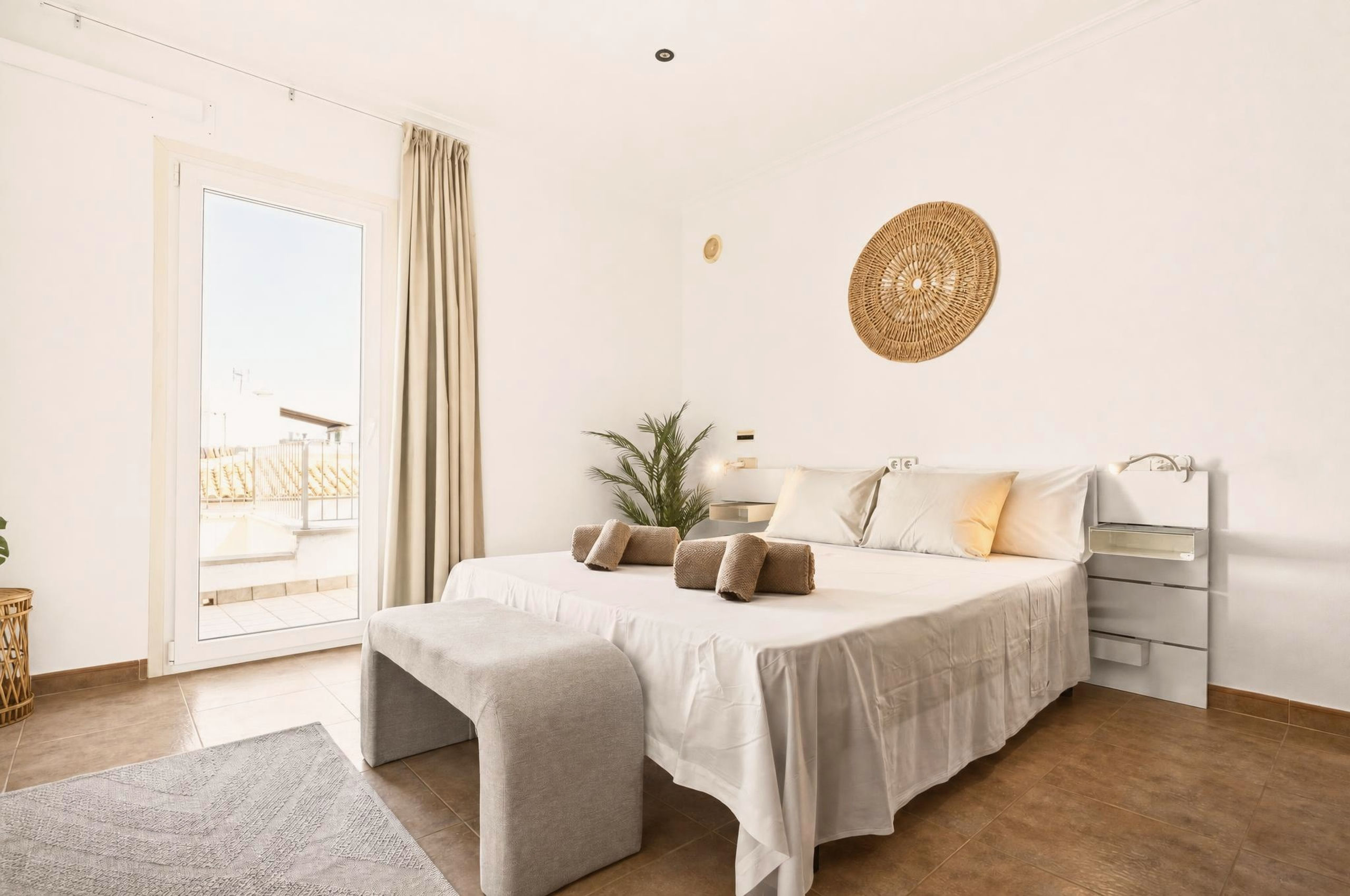 Helles Doppelschlafzimmer mit beigem Leinenbett, gerollten Handtüchern, grauer Bank, Rattan-Wandschmuck und Glastür zur sonnigen Terrasse.