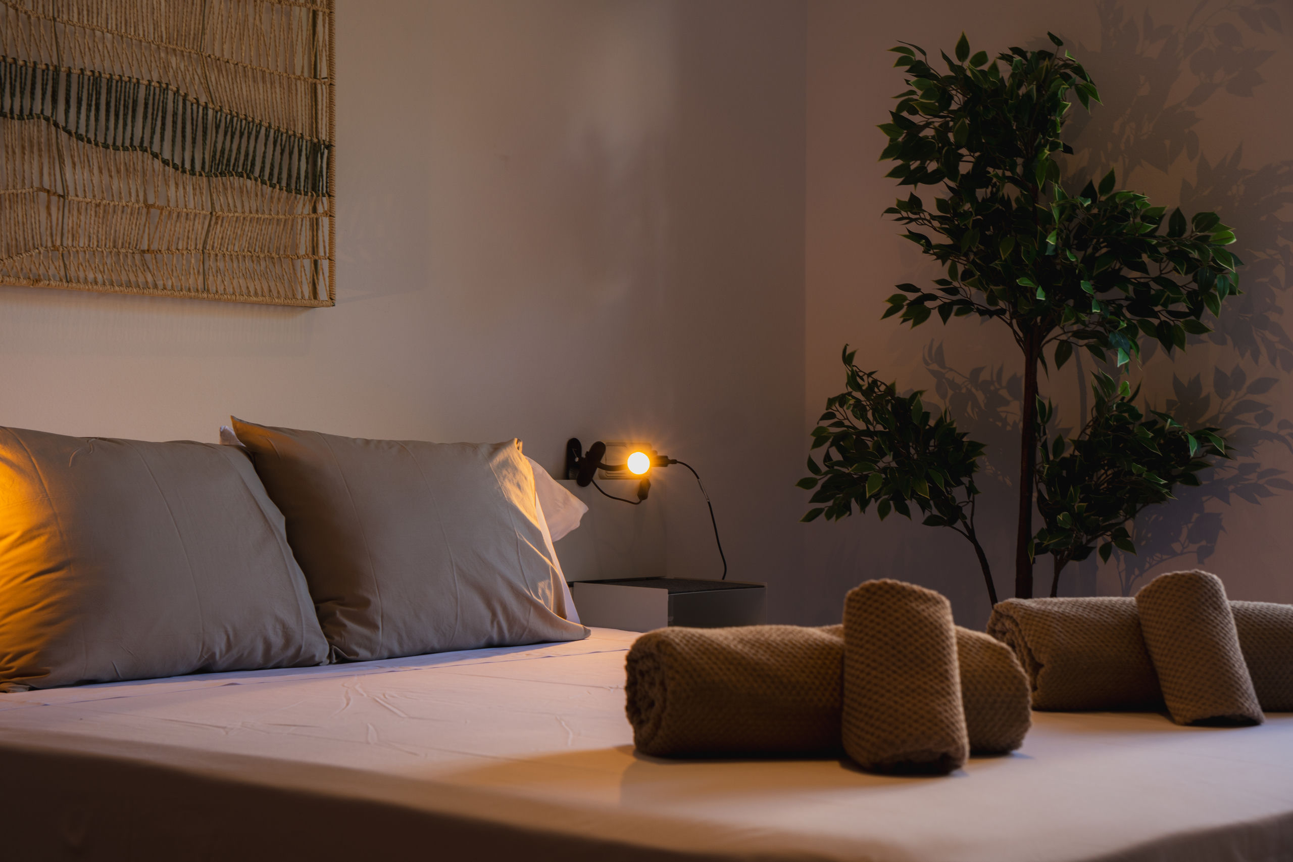 Gedämpfte abendliche Szene im selben Zimmer: warme Leselampe an grauen Kissen, Blattpflanze rechts, gerollte Handtücher auf dem Bett.