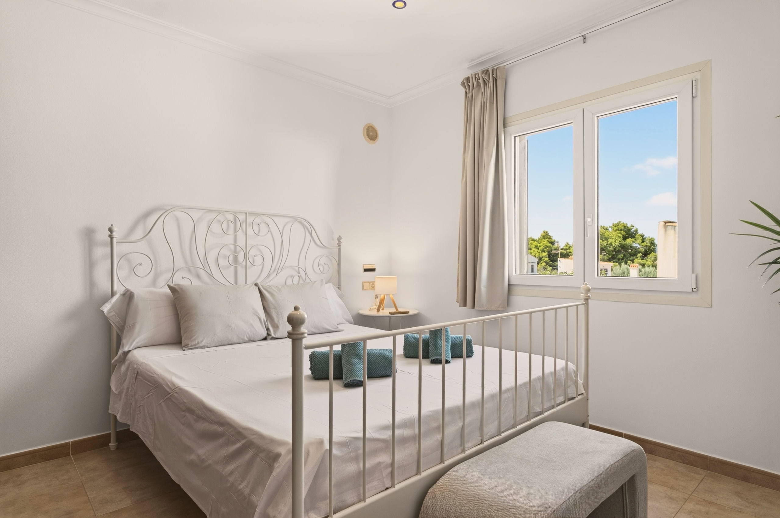 Drittes Schlafzimmer mit weißem Metallbett mit verziertem Kopfteil, beiger Bettwäsche, türkisfarbenen Handtüchern, leuchtender Dreifußlampe und großem Fenster.