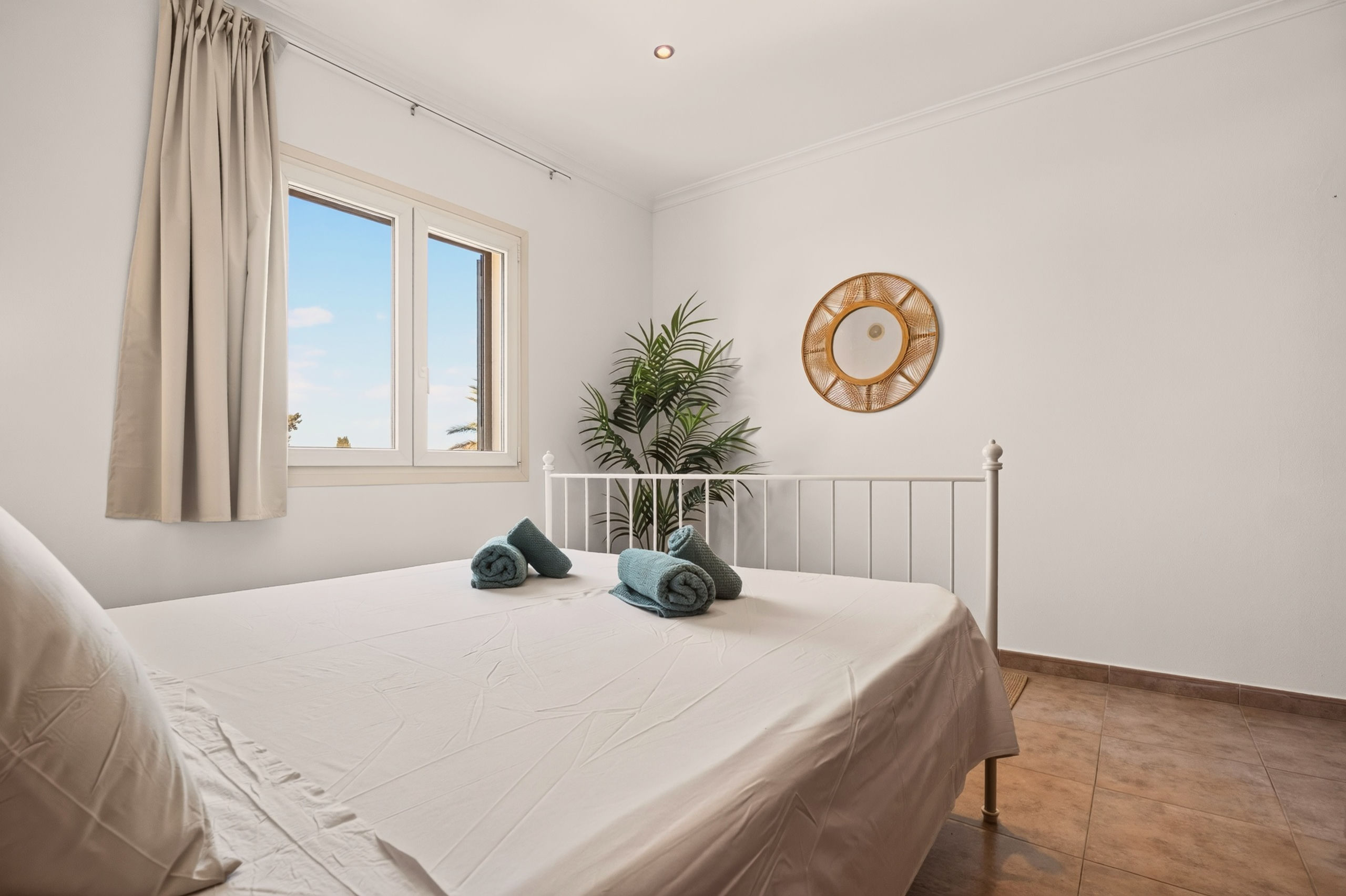 Gegenüberliegende Eckansicht: Metallbett mit türkisen Handtüchern, runder Rattanspiegel und Palme hinter dem Fußteil, Fenster mit Himmel links.