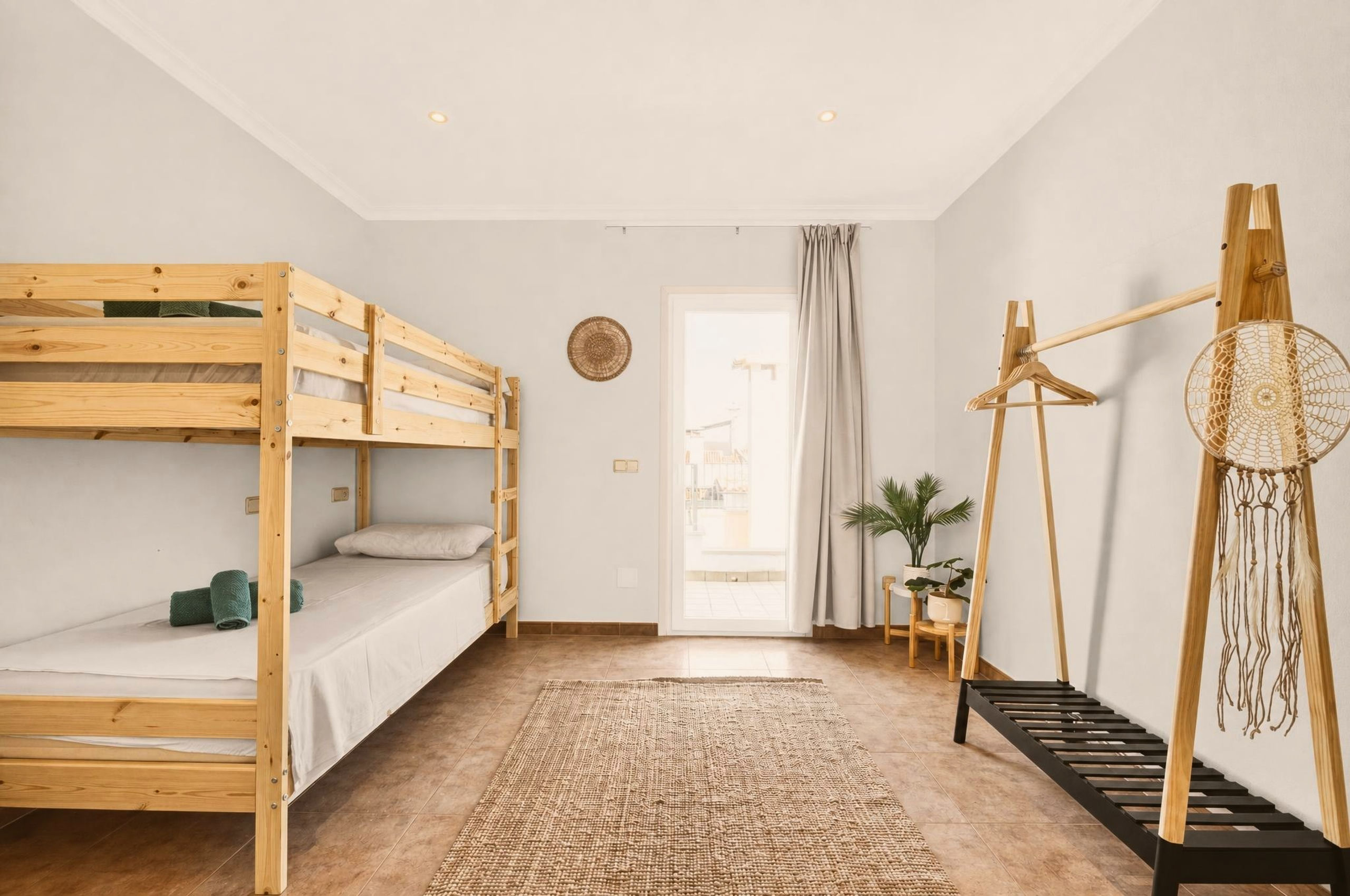 Etagenbettzimmer mit Bettgestell aus Kiefernholz, Jute-Teppich, Kleiderständer aus Holz mit Traumfänger und Glastür zur hellen Terrasse.