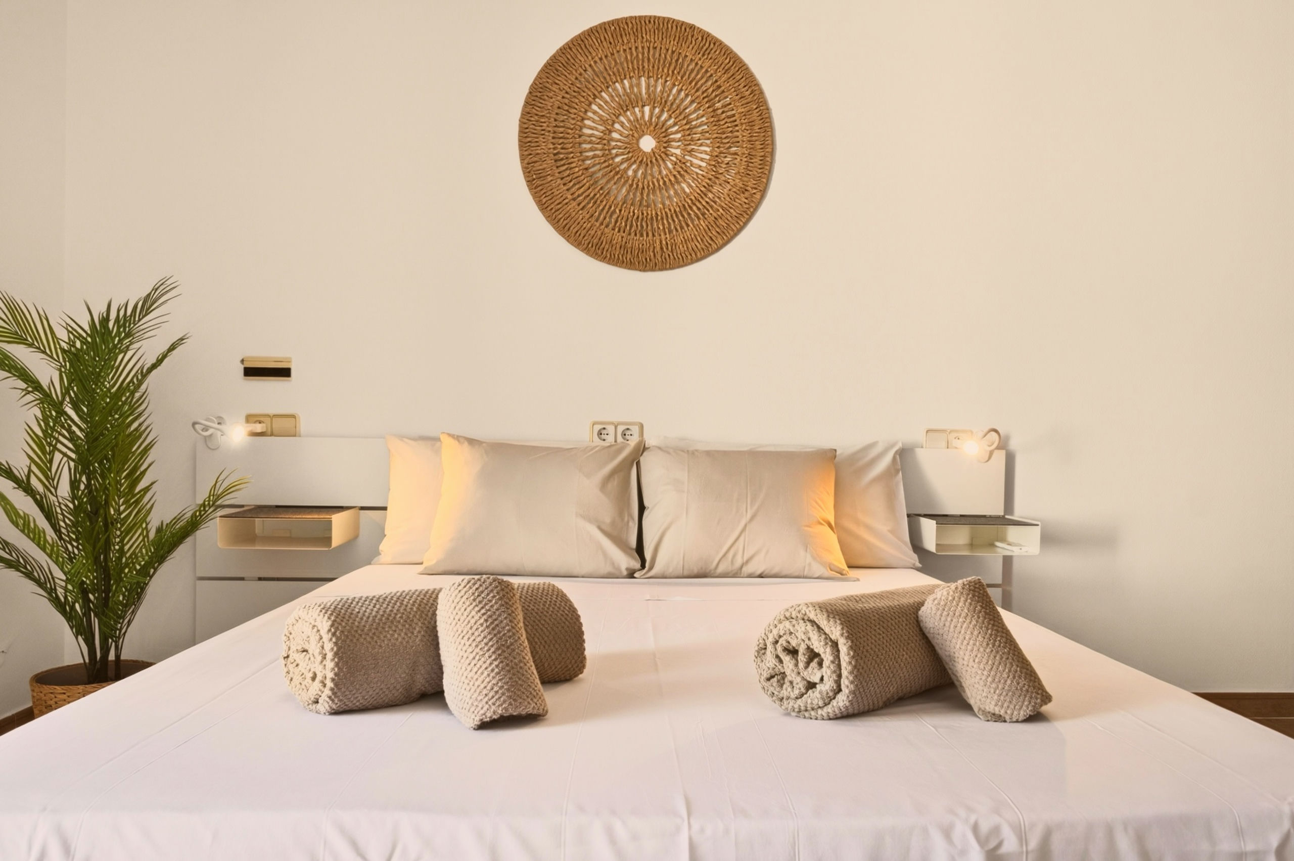 Frontalansicht des gemachten Betts: beige Kissen, vier gerollte Handtücher, weiße Schwebe-Nachttische mit Leselampen und Rattan-Mandala darüber.
